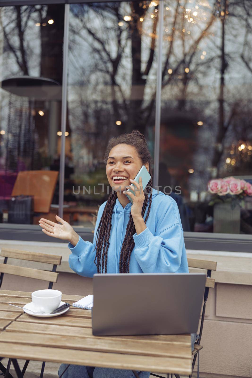 Pretty smiled young mixed-race female talking on cellphone while having coffee outdoor at laptop. by Yaroslav_astakhov