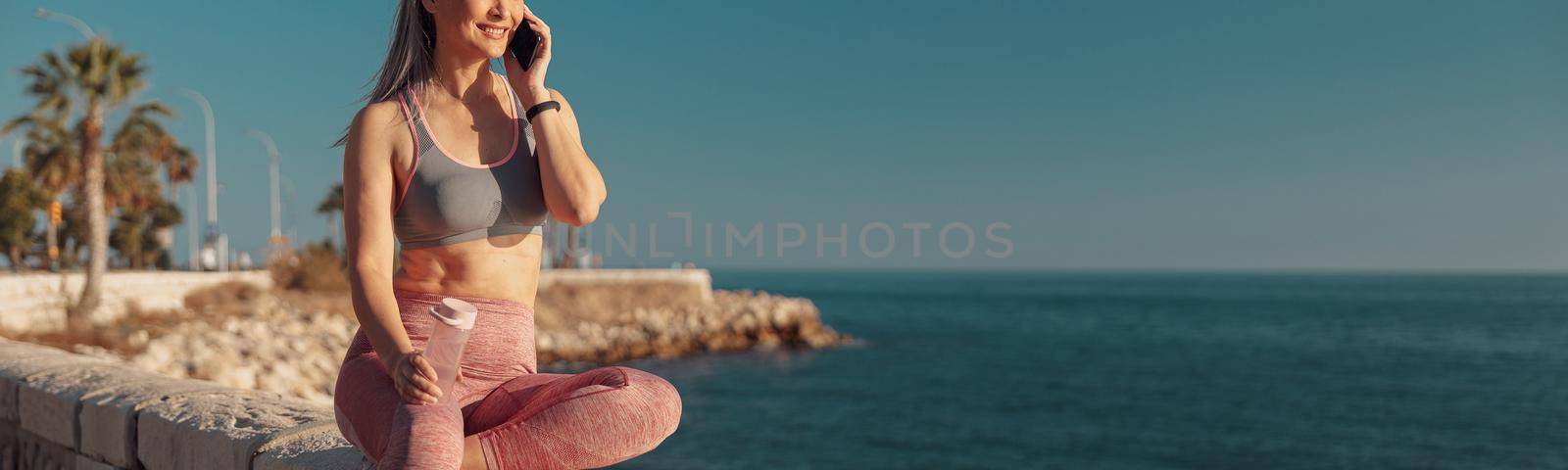 Smiling woman in sports clothes sitting by the seashore, talking on the phone and looking at the sun