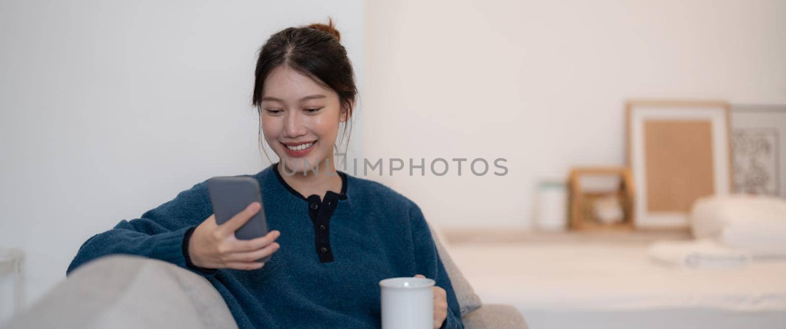 Happy young asian woman using mobile phone while sitting a couch at home.