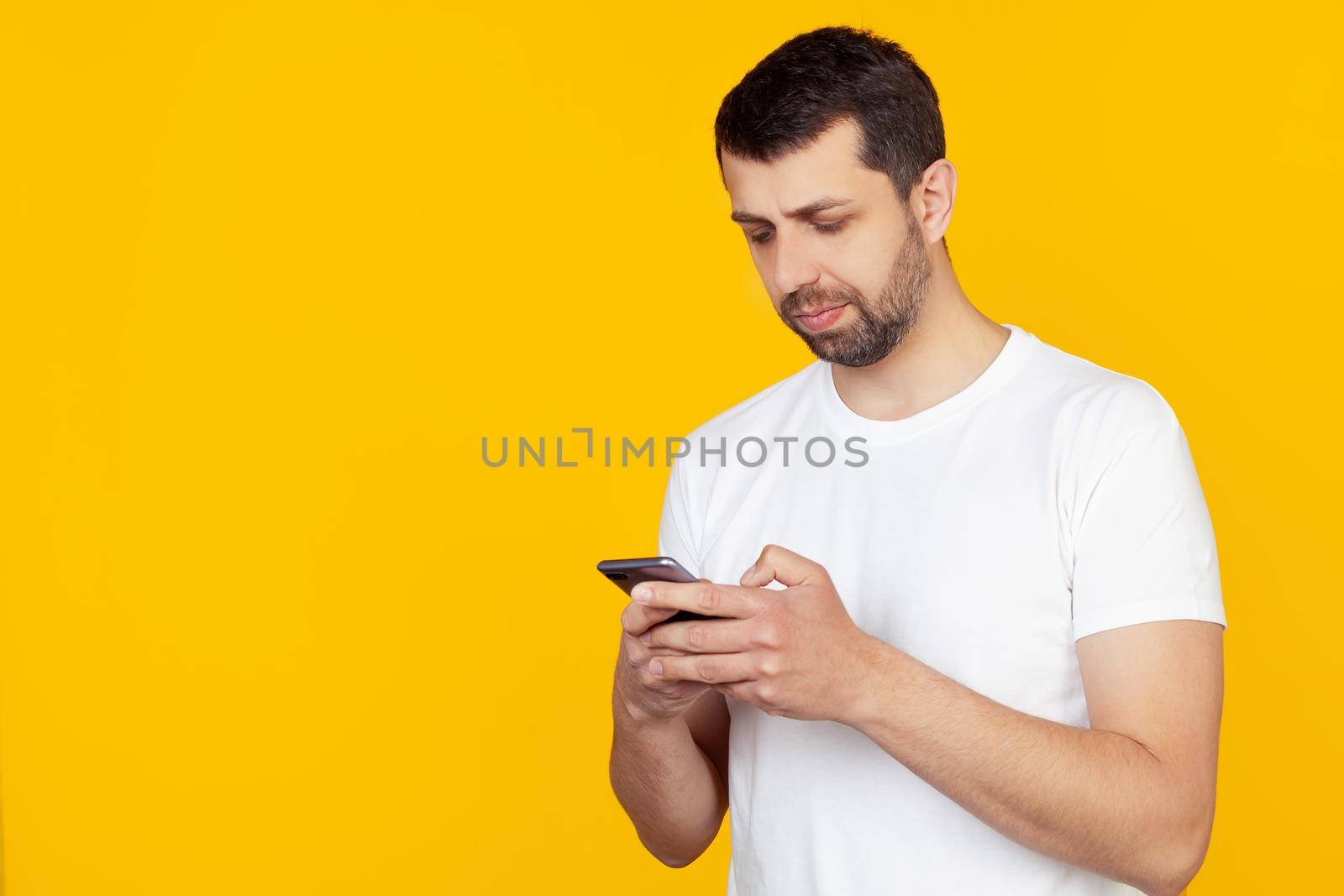 A young man with a beard in a white t-shirt uses a smartphone with a confident expression on his smart face, thinking seriously. Stands on isolated yellow background.