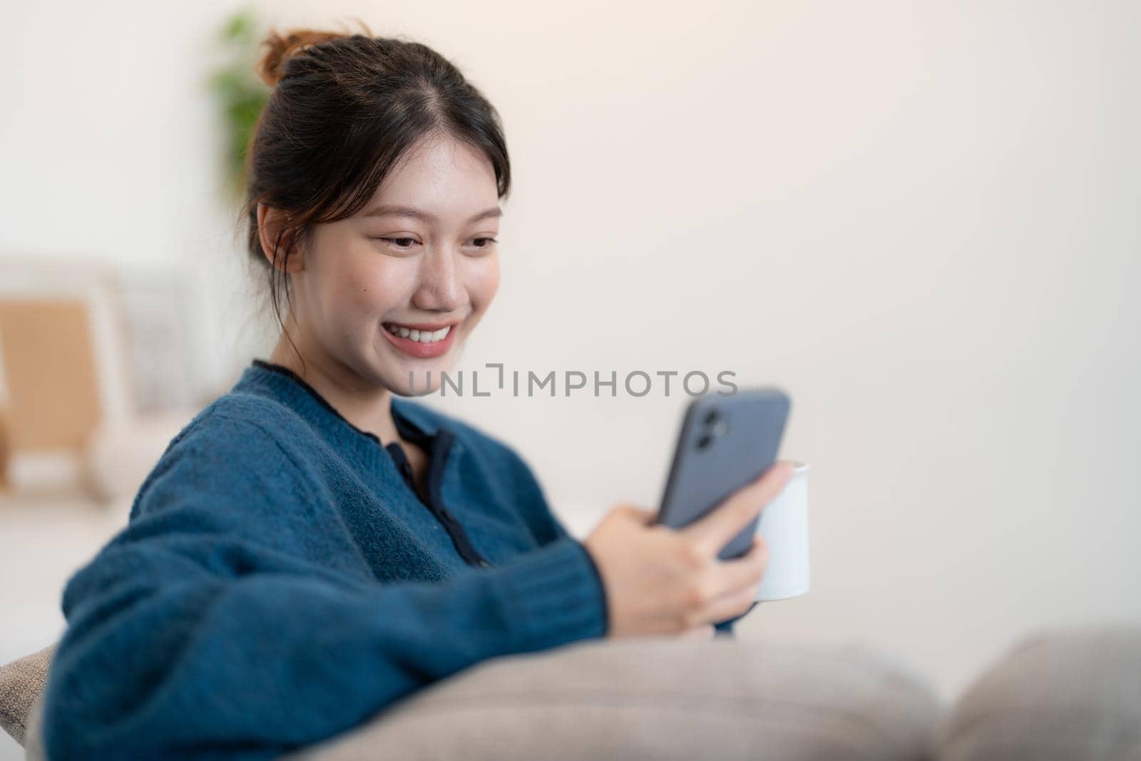 Happy young asian woman using mobile phone while sitting a couch at home.