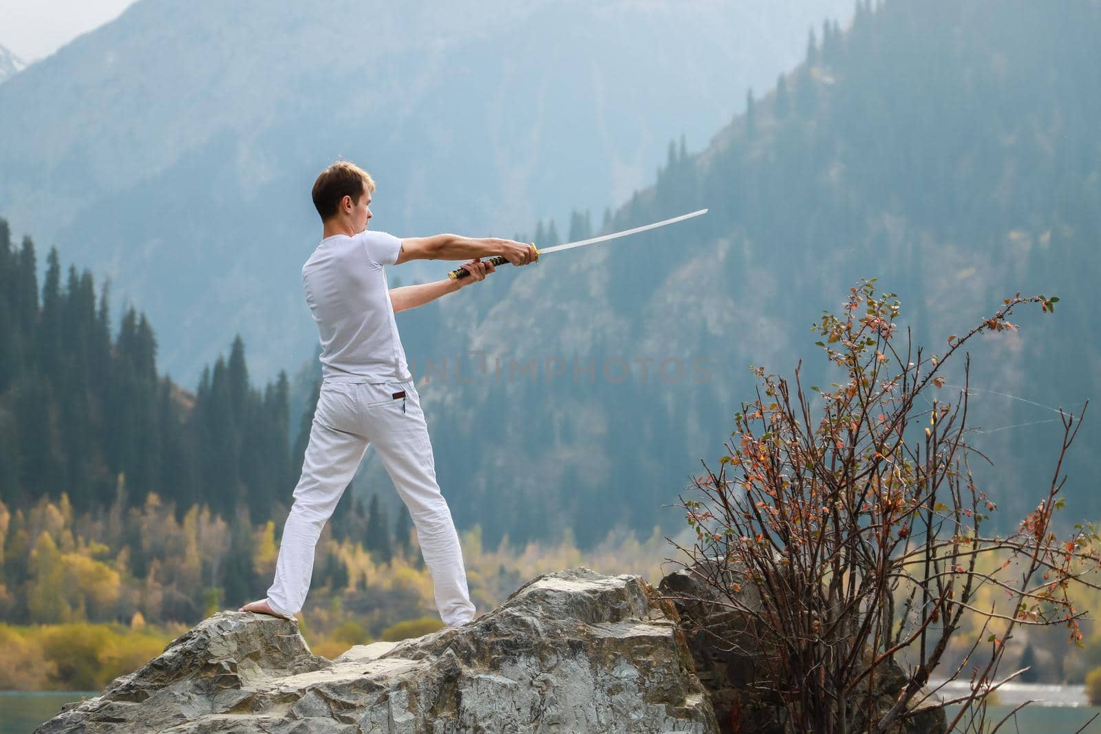 A young man stands on a large stone and holds a Japanese sword in his hands