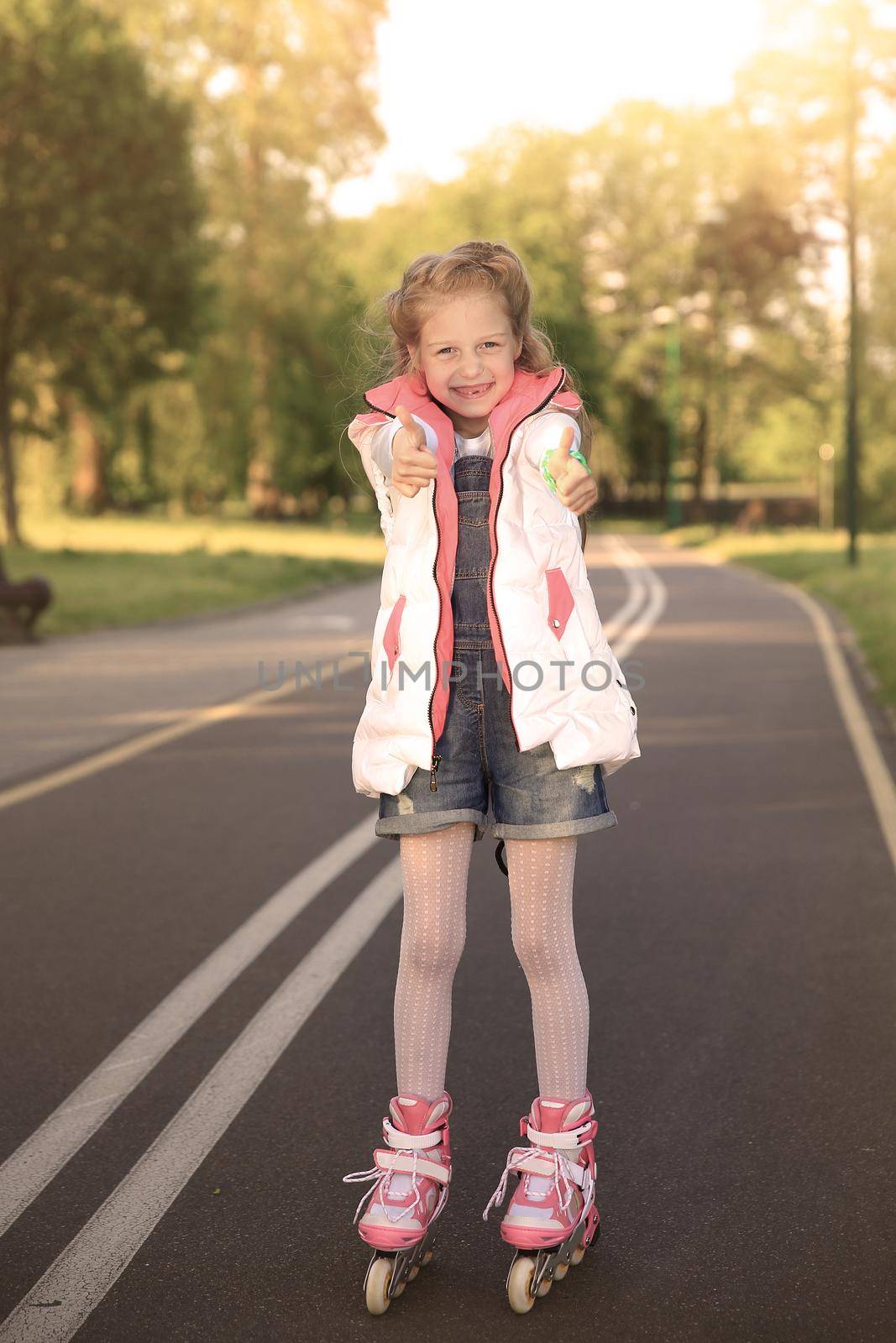 little girl on roller skates makes a gesture thumb up in the Park on a Sunny day