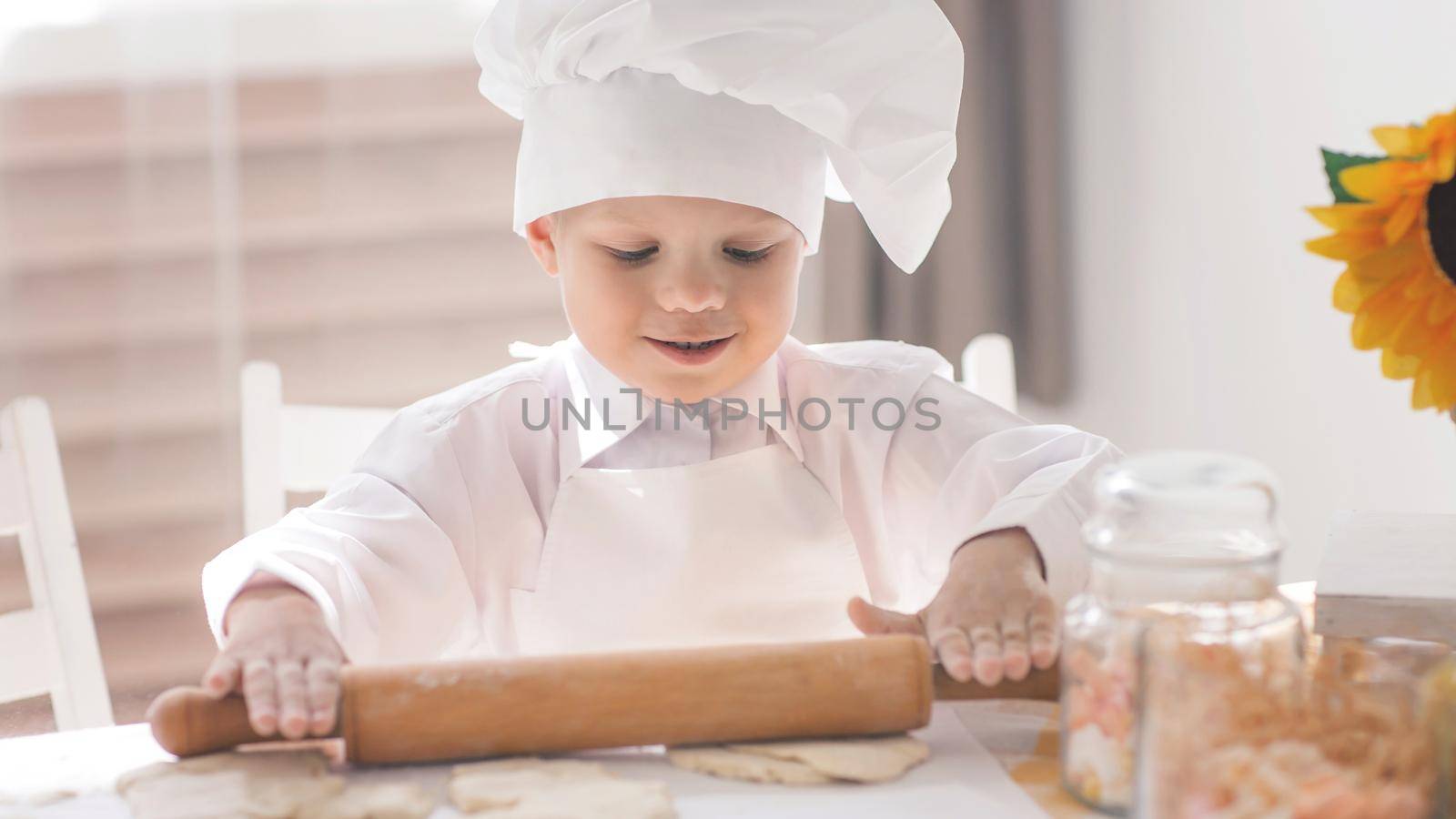 A small child in the form of a cook rolls out the dough on the kitchen table