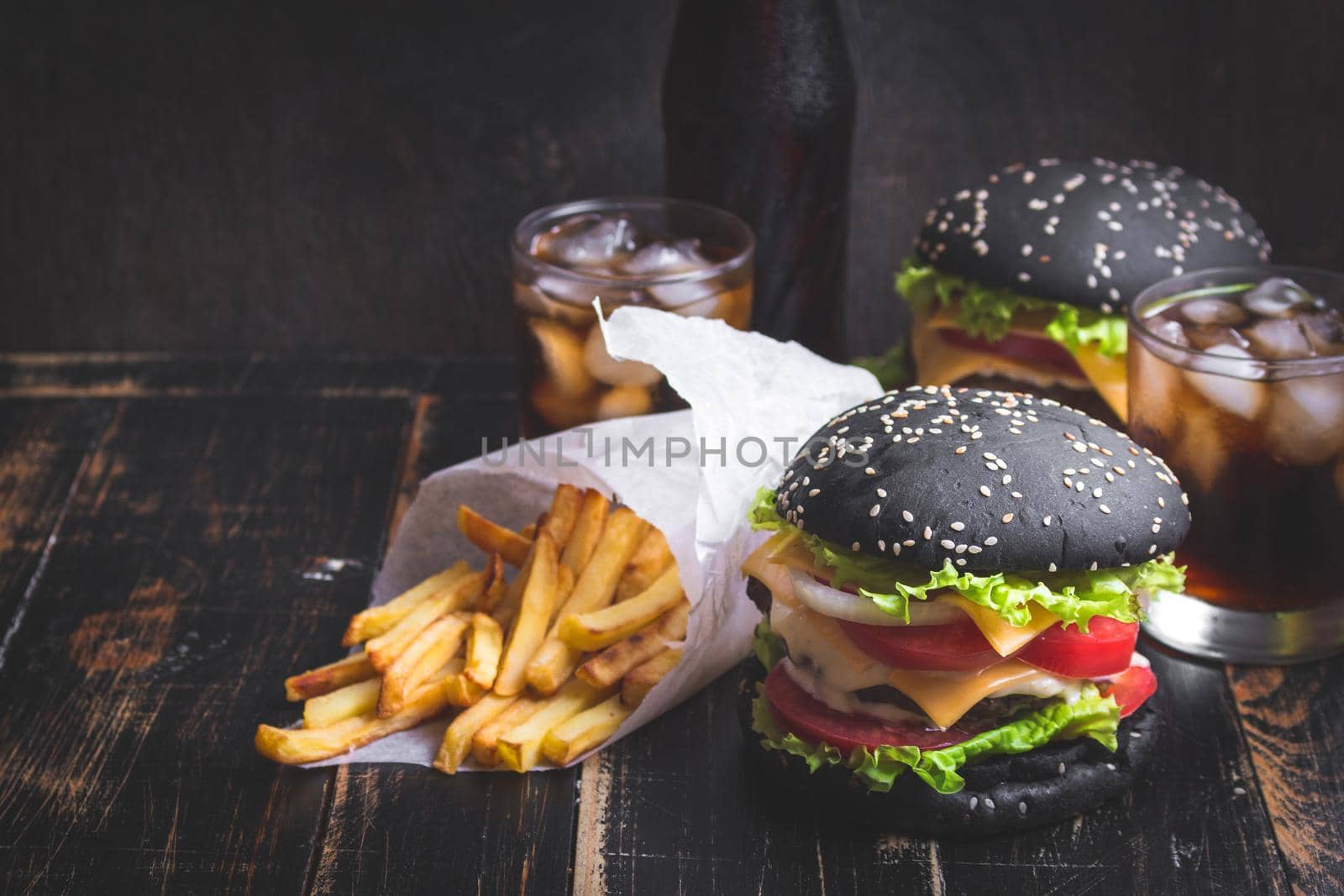 Set of black burgers with meat patty, cheese, tomatoes, mayonnaise, french fries in a paper cup and glass of cold cola soda with ice. Dark wooden rustic table. Modern fast food lunch. Toned image