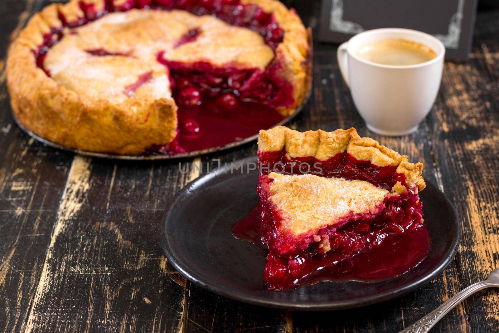Slice of homemade cherry pie, cup of coffee, bowl with cherries and menu chalkboard on the black wooden table