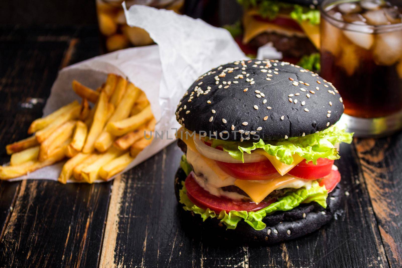 Black burger with meat patty, cheese, tomatoes, mayonnaise, french fries in a paper cup and glass of cold cola soda with ice. Dark wooden rustic table. Modern fast food lunch