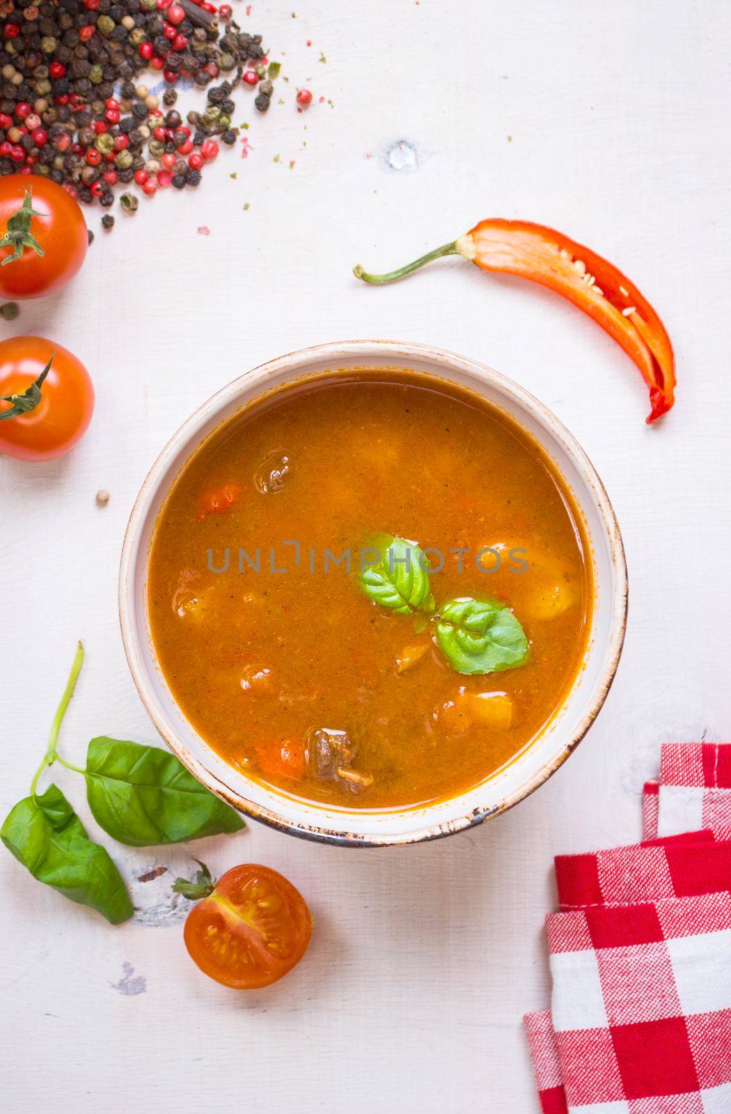 Delicious tomato soup with meat in a white bowl on a wooden table with fresh cherry tomatoes, basil leaves, cut chili pepper and red gingham kitchen towel. Ingredients for soup. Top view