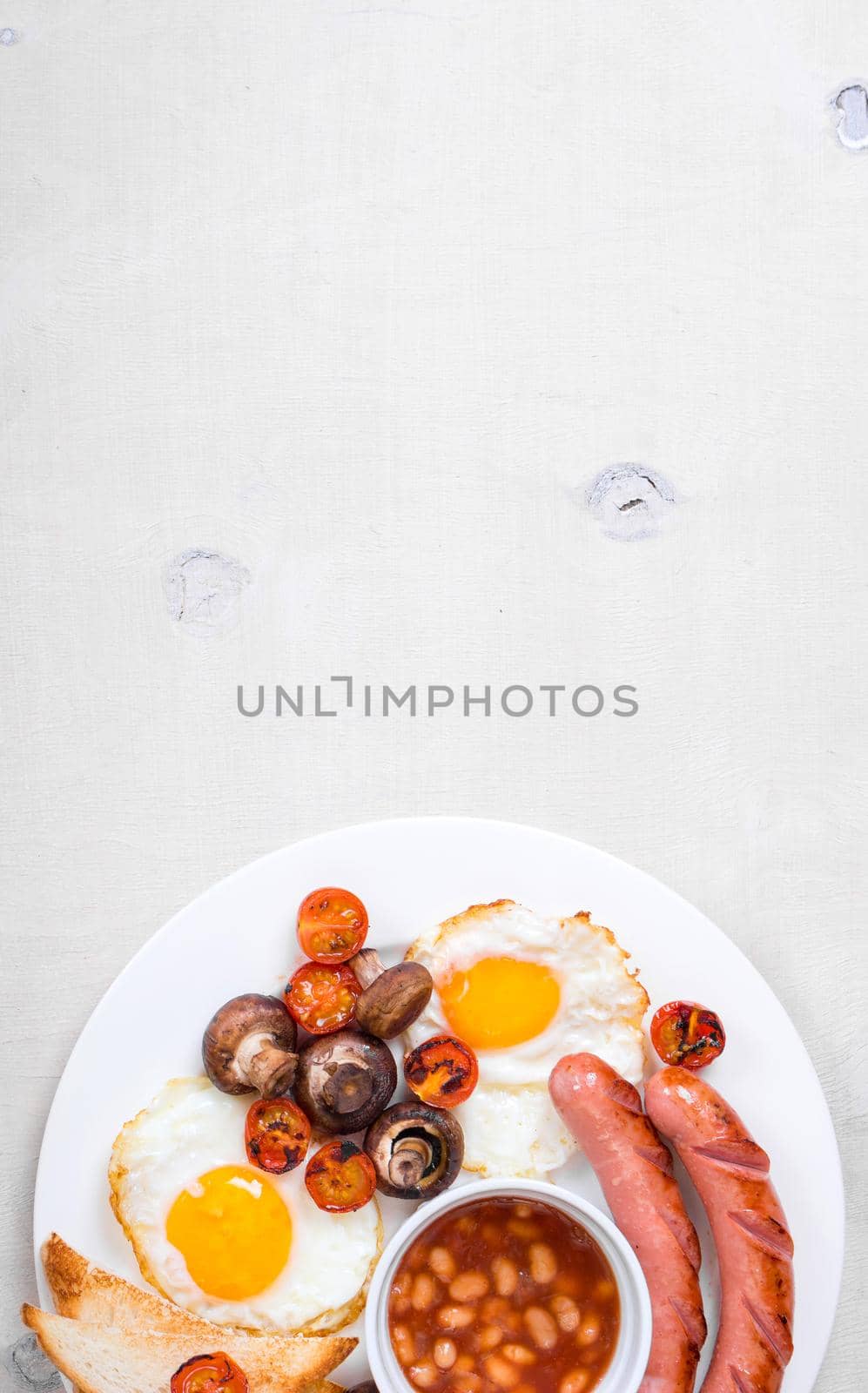 Full english breakfast with fried eggs, tomatoes, sausages, bacon, mushrooms, toasts and beans. Breakfast on a white plate with forks on the white wooden table. Space for text. Background