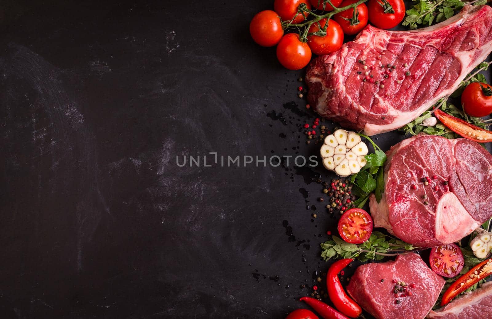 Raw juicy meat steaks ready for roasting on a black chalk board background. Rib eye steak on the bone, veal shank (ossobuco), fillet with cherry tomatoes, hot pepper and herbs. Space for text