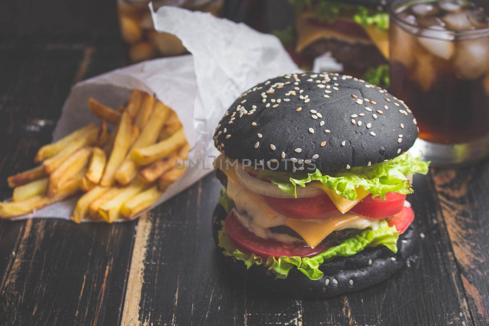 Black burger with meat patty, cheese, tomatoes, mayonnaise, french fries in a paper cup and glass of cold cola soda with ice. Dark wooden rustic table. Modern fast food lunch. Toned image