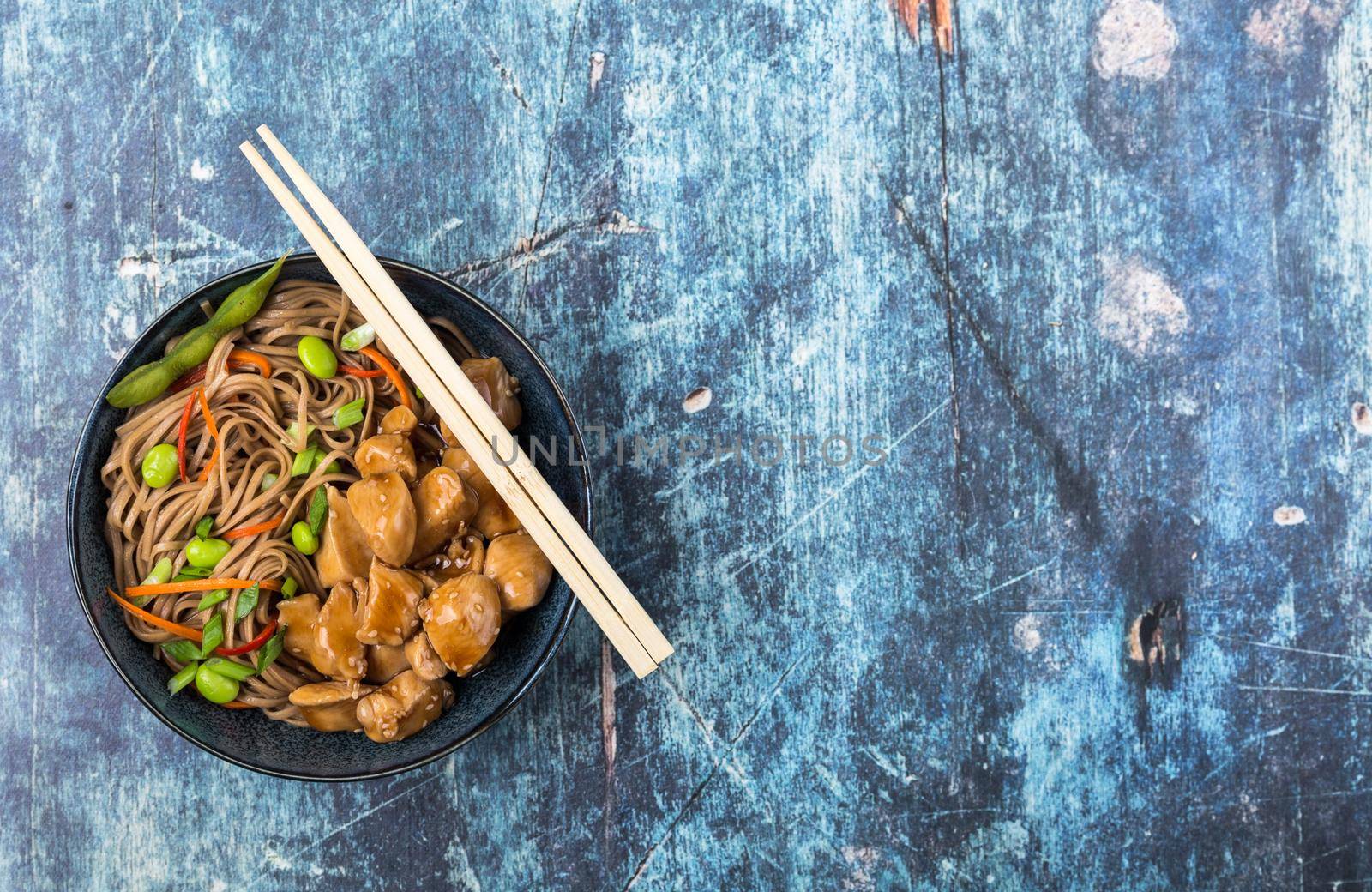 Asian noodles with chicken, vegetables, bowl, rustic wooden blue background. Space for text. Top view. Soba noodles, teriyaki chicken, edamame, chopsticks. Asian style dinner. Chinese/Japanese noodles