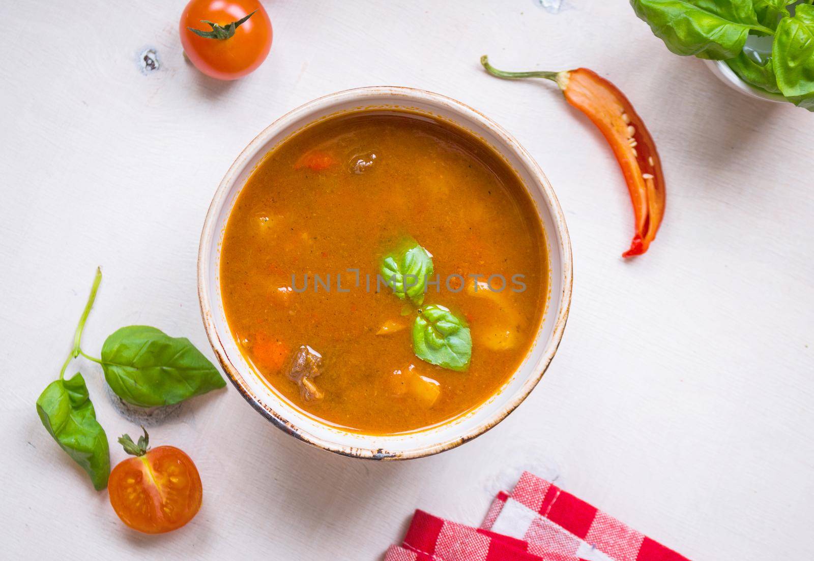 Delicious tomato soup with meat in a white bowl on a wooden table with fresh cherry tomatoes, basil leaves, cut chili pepper and red gingham kitchen towel. Ingredients for soup. Top view