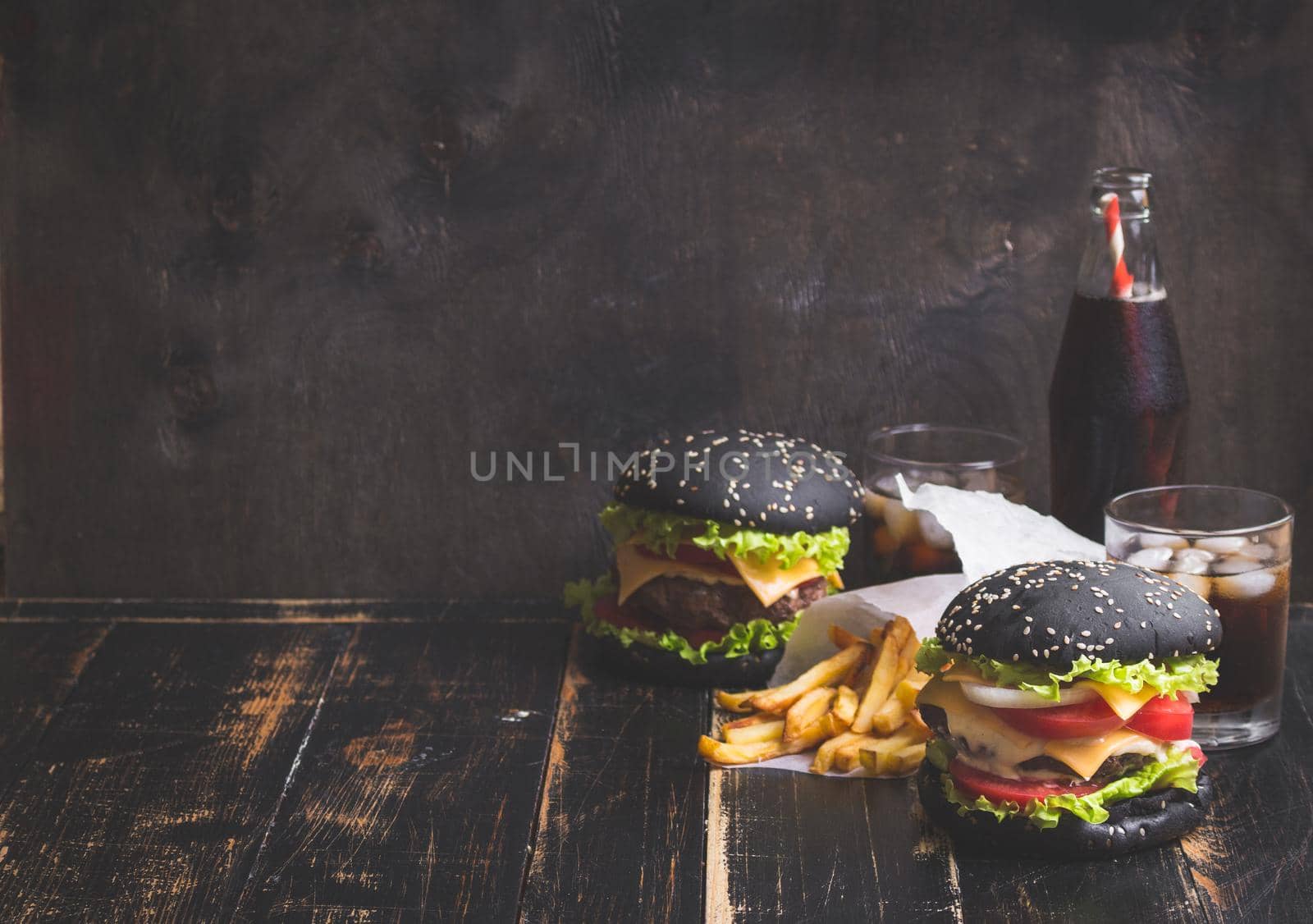 Set of black burgers with meat patty, cheese, tomatoes, mayonnaise, french fries and glass of cold cola soda with ice. Dark wooden rustic background. Space for text. Modern fast food. Toned image