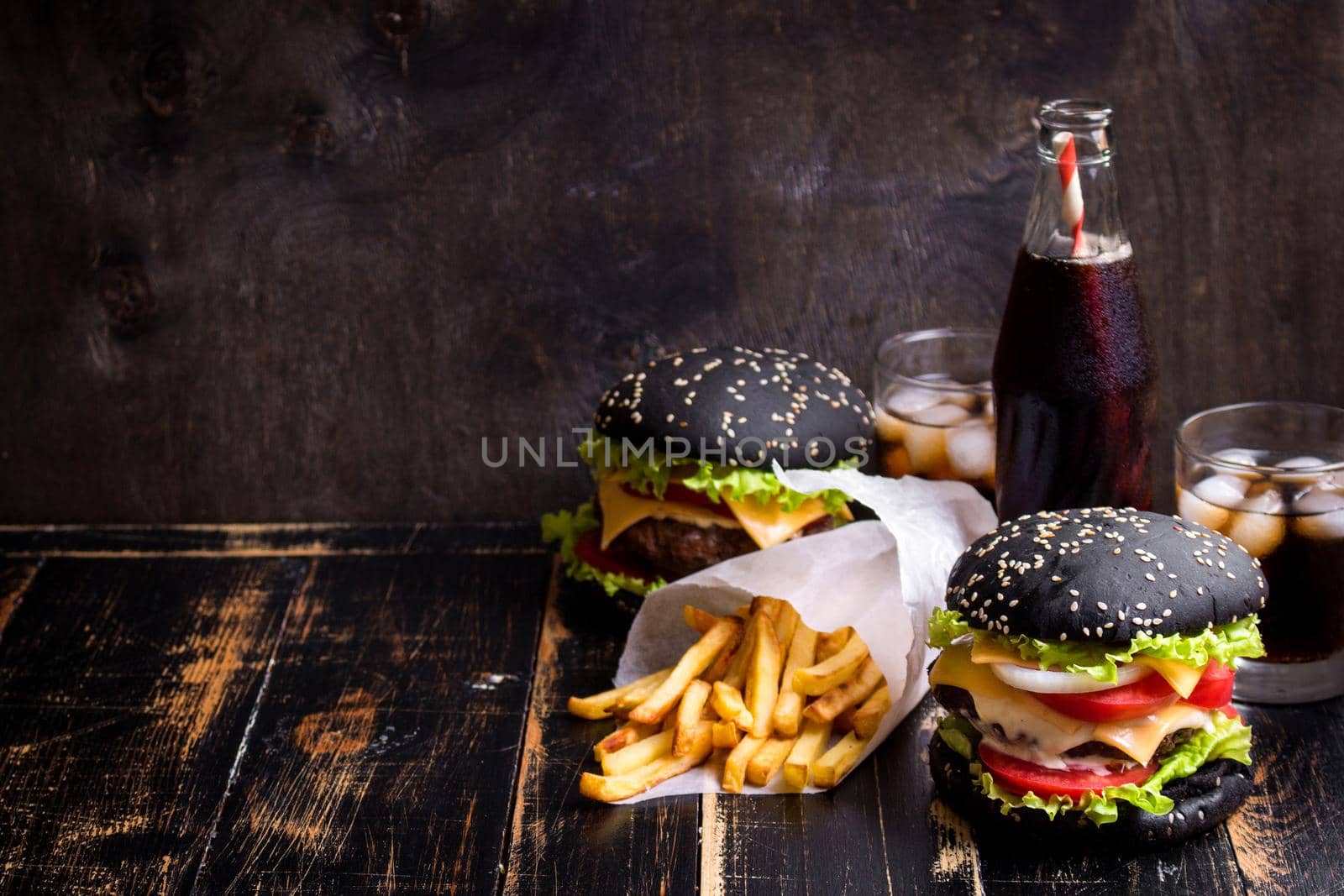 Set of black burgers with meat patty, cheese, tomatoes, mayonnaise, french fries and glass of cold cola soda with ice. Dark wooden rustic background. Space for text. Modern fast food lunch frame