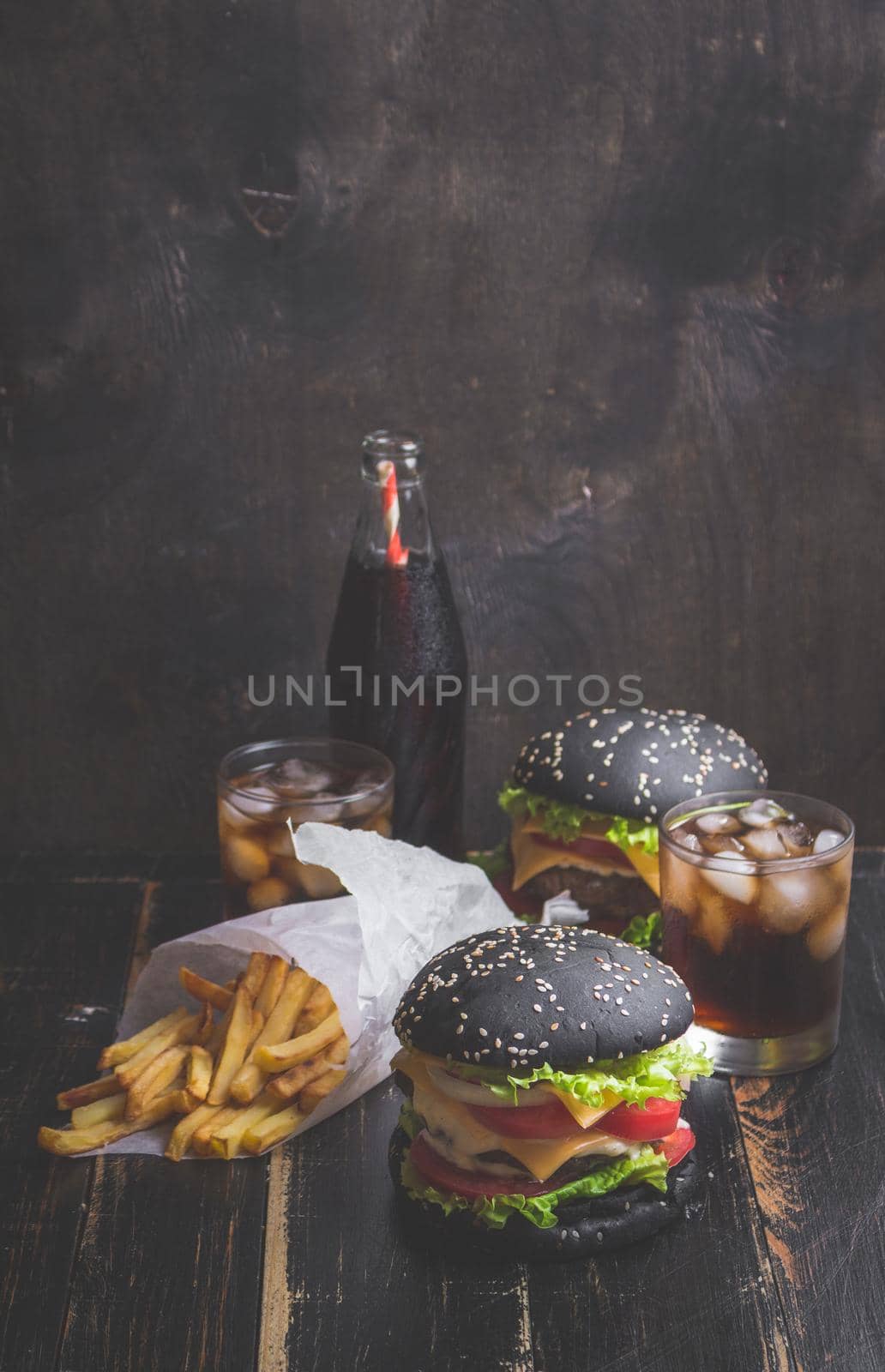Set of black burgers with meat patty, cheese, tomatoes, mayonnaise, french fries in a paper cup and glass of cold cola soda with ice. Dark wooden rustic table. Modern fast food lunch. Toned image