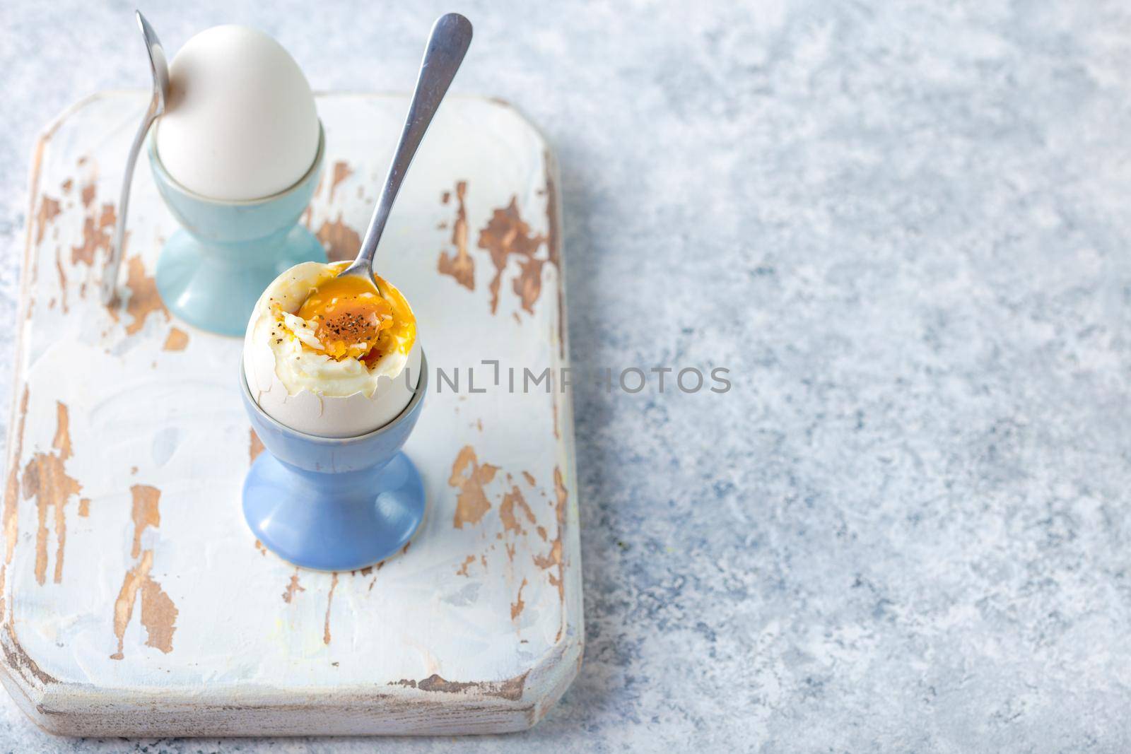 Fresh soft boiled eggs in stands, spoons, white wooden cooking board, white concrete rustic background. Soft eggs for healthy breakfast. Space for text. Selective focus. Egg protein fitness breakfast