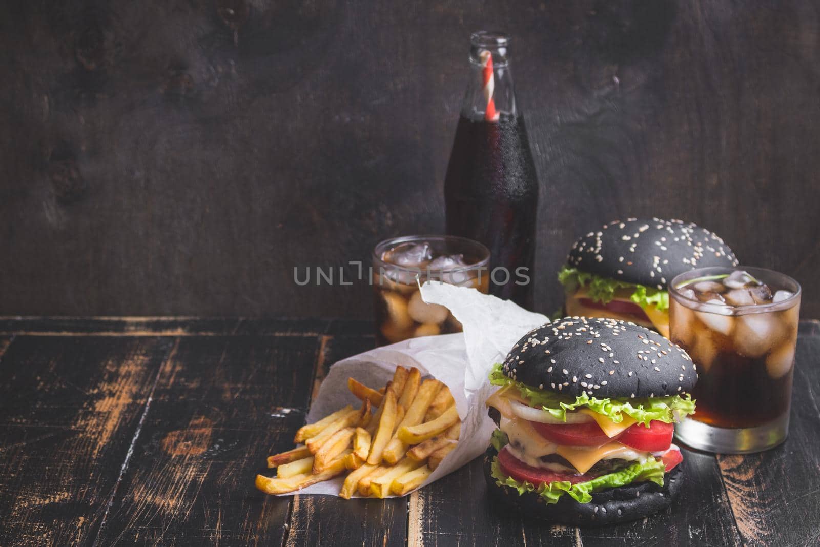 Set of black burgers with meat patty, cheese, tomatoes, mayonnaise, french fries and glass of cold cola soda with ice. Dark wooden rustic background. Space for text. Modern fast food. Toned image