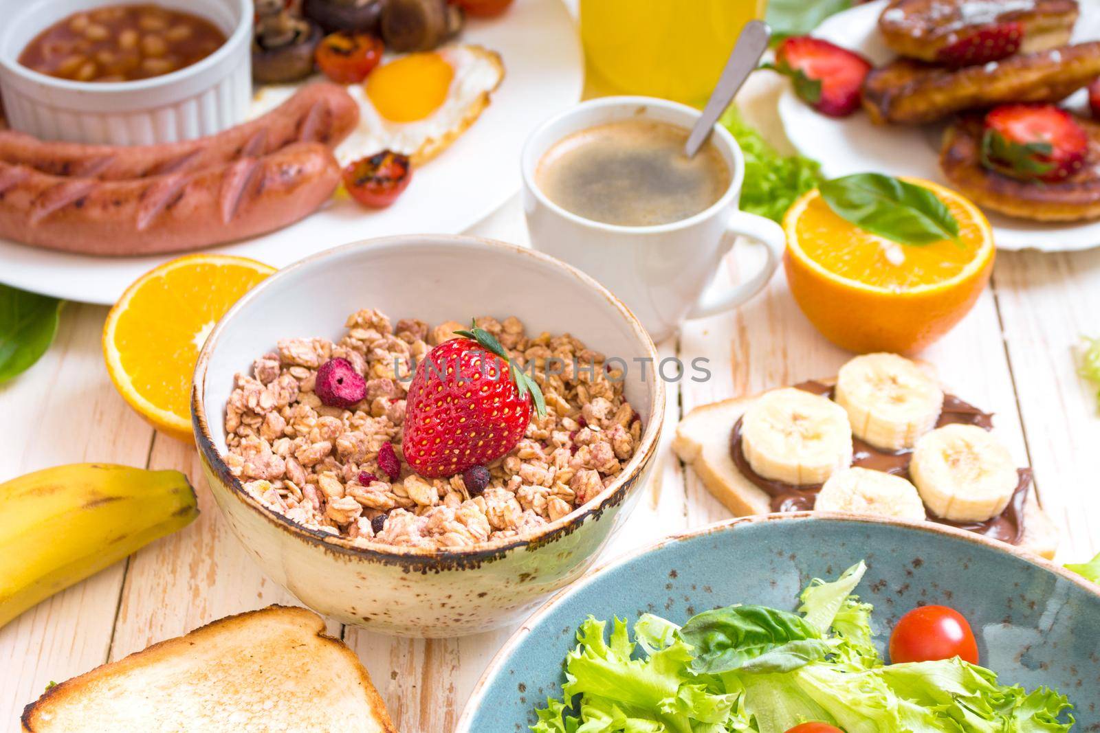 Assortment of breakfast choices. Granola with strawberry in a bowl and sausages, fried eggs, bacon, salad, cheese sandwich, pancakes, chocolate cream and banana toast, coffee, orange juice. Close-up