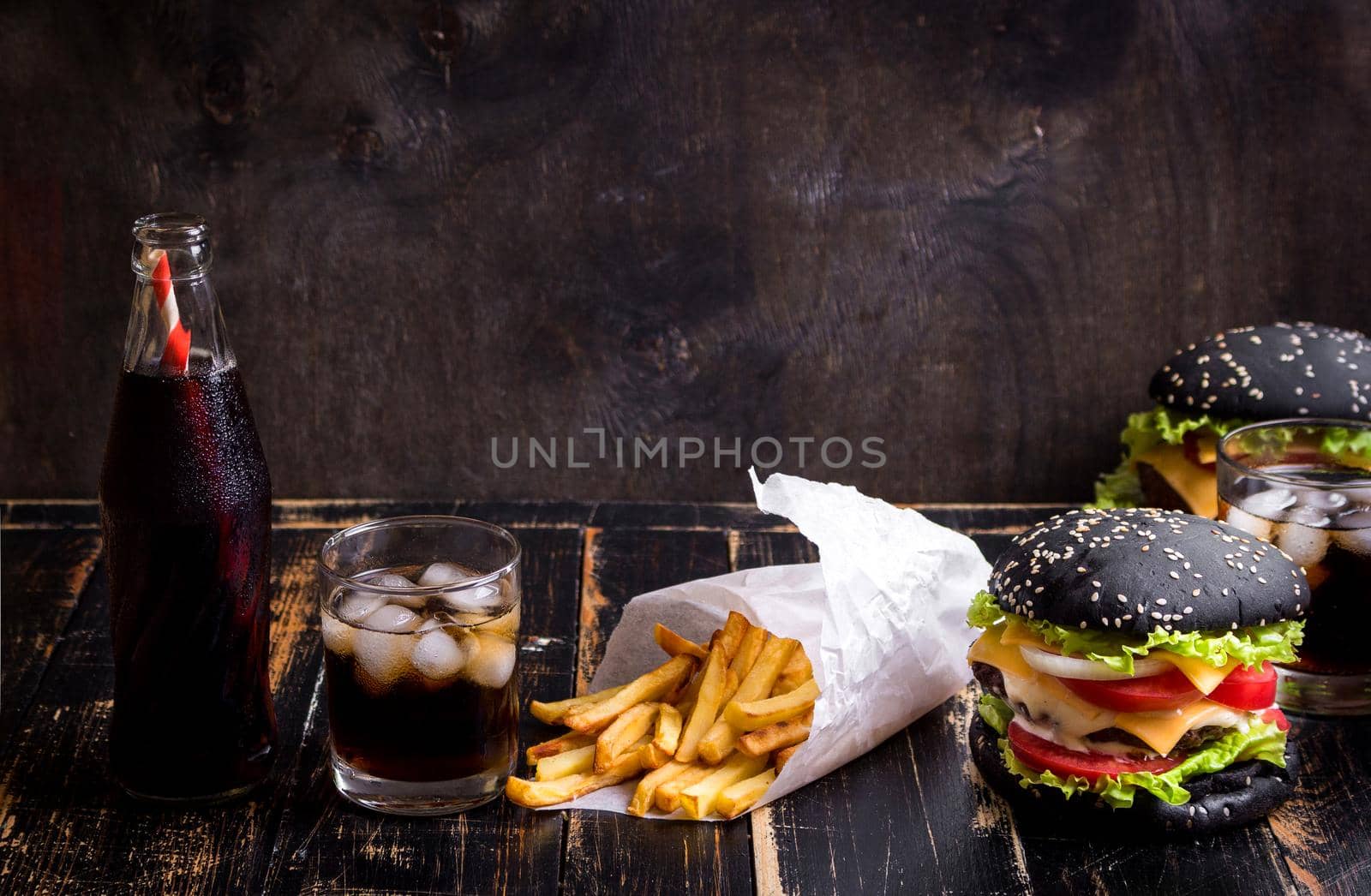 Set of black burgers with meat patty, cheese, tomatoes, mayonnaise, french fries and glass of cold cola soda with ice. Dark wooden rustic background. Space for text. Modern fast food lunch frame