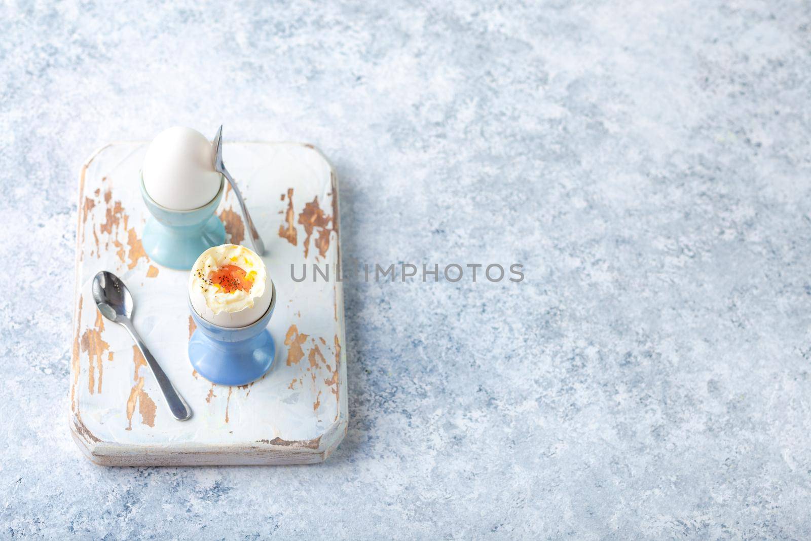 Fresh soft boiled eggs in stands, spoons, wooden cooking board, white concrete rustic background. Top view. Soft eggs, healthy breakfast. Space for text. Selective focus. Egg protein fitness breakfast