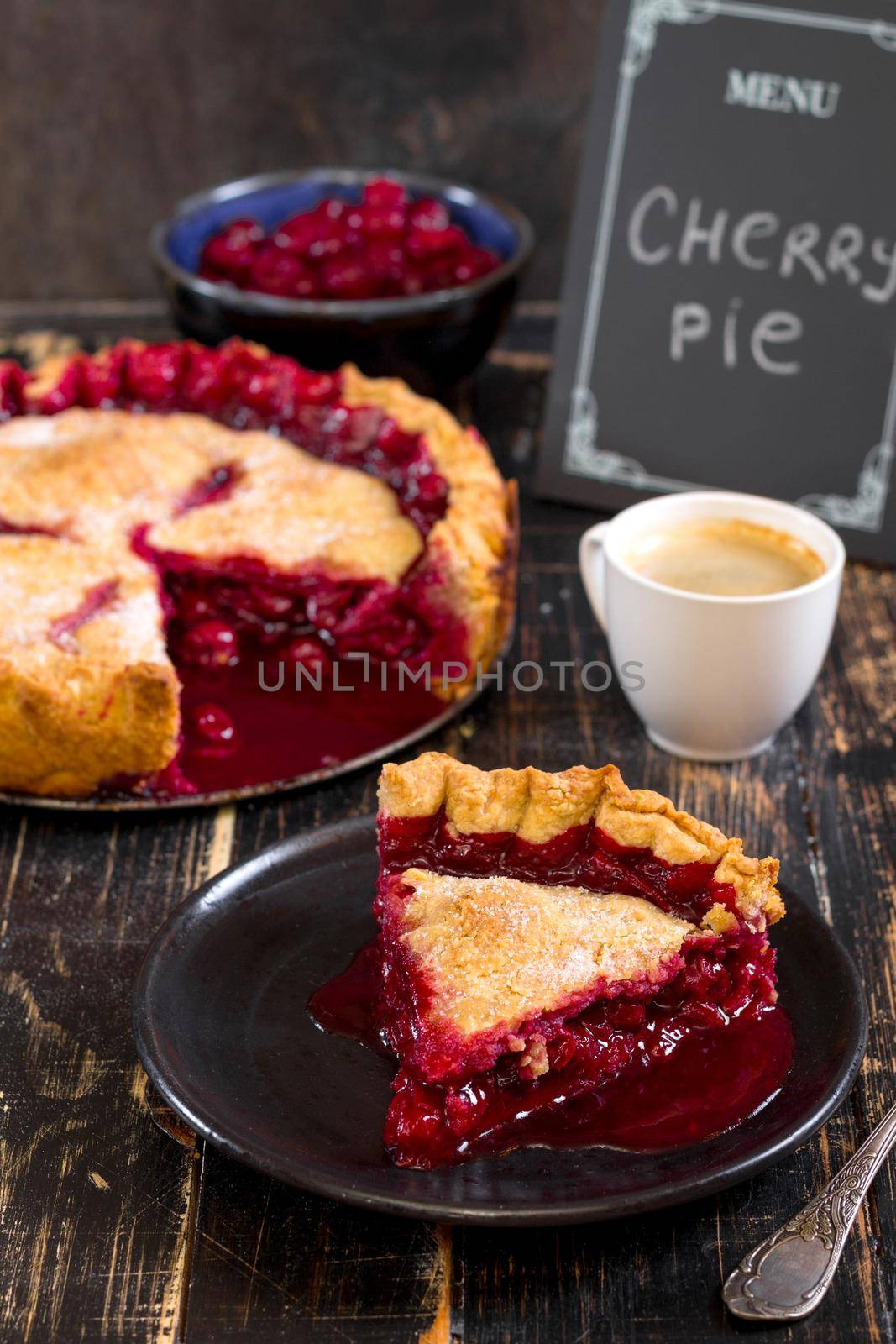 Slice of homemade cherry pie, cup of coffee, bowl with cherries and menu chalkboard on the black wooden table