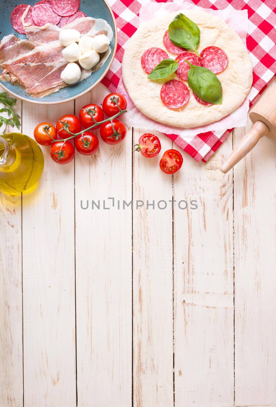 Pizza making background. Ingredients for making pizza. Space for text. Pizza dough, flour, cheese, mozzarella, tomatoes, basil, pepperoni, olives and rolling pin over white wooden background. Top view