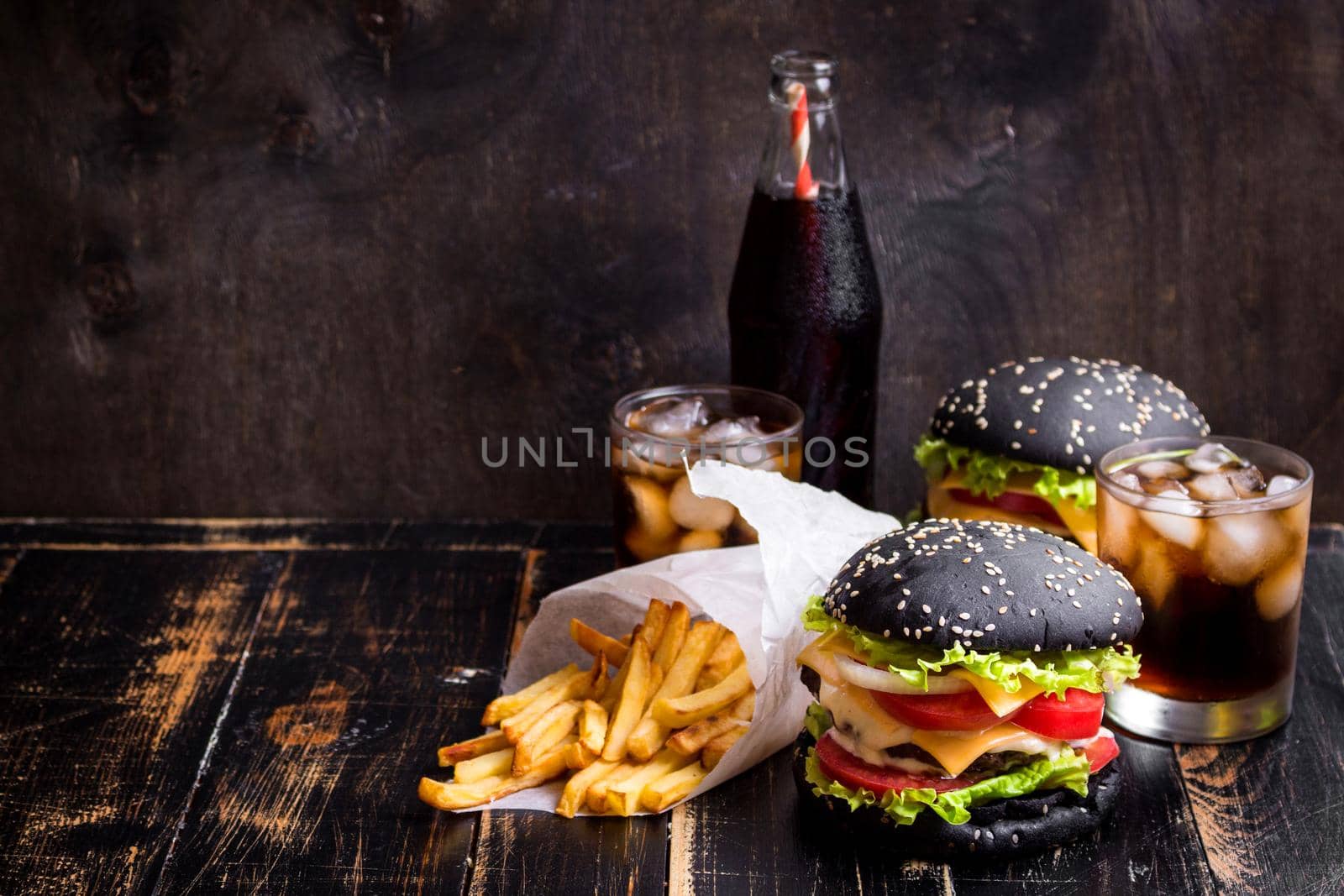 Set of black burgers with meat patty, cheese, tomatoes, mayonnaise, french fries and glass of cold cola soda with ice. Dark wooden rustic background. Space for text. Modern fast food lunch frame