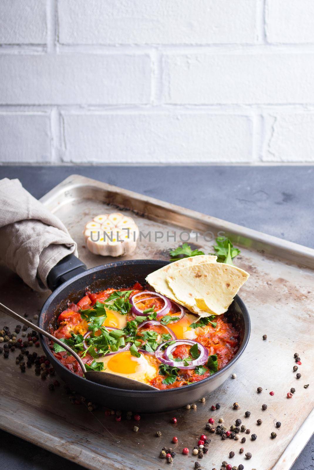 Shakshuka with pita bread in a pan. Middle eastern traditional dish. Fried eggs with tomatoes, bell pepper, vegetables and herbs. Shakshouka on a table. Sunny side up eggs. Homemade. Selective focus