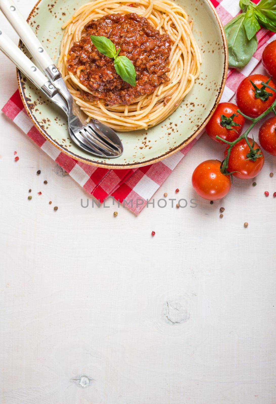 Italian pasta bolognese. Spaghetti with meat and tomato sauce in a plate with Italian tablecloth on a wooden white background. With fresh cherry tomatoes, basil. Food frame. Space for text. Top view