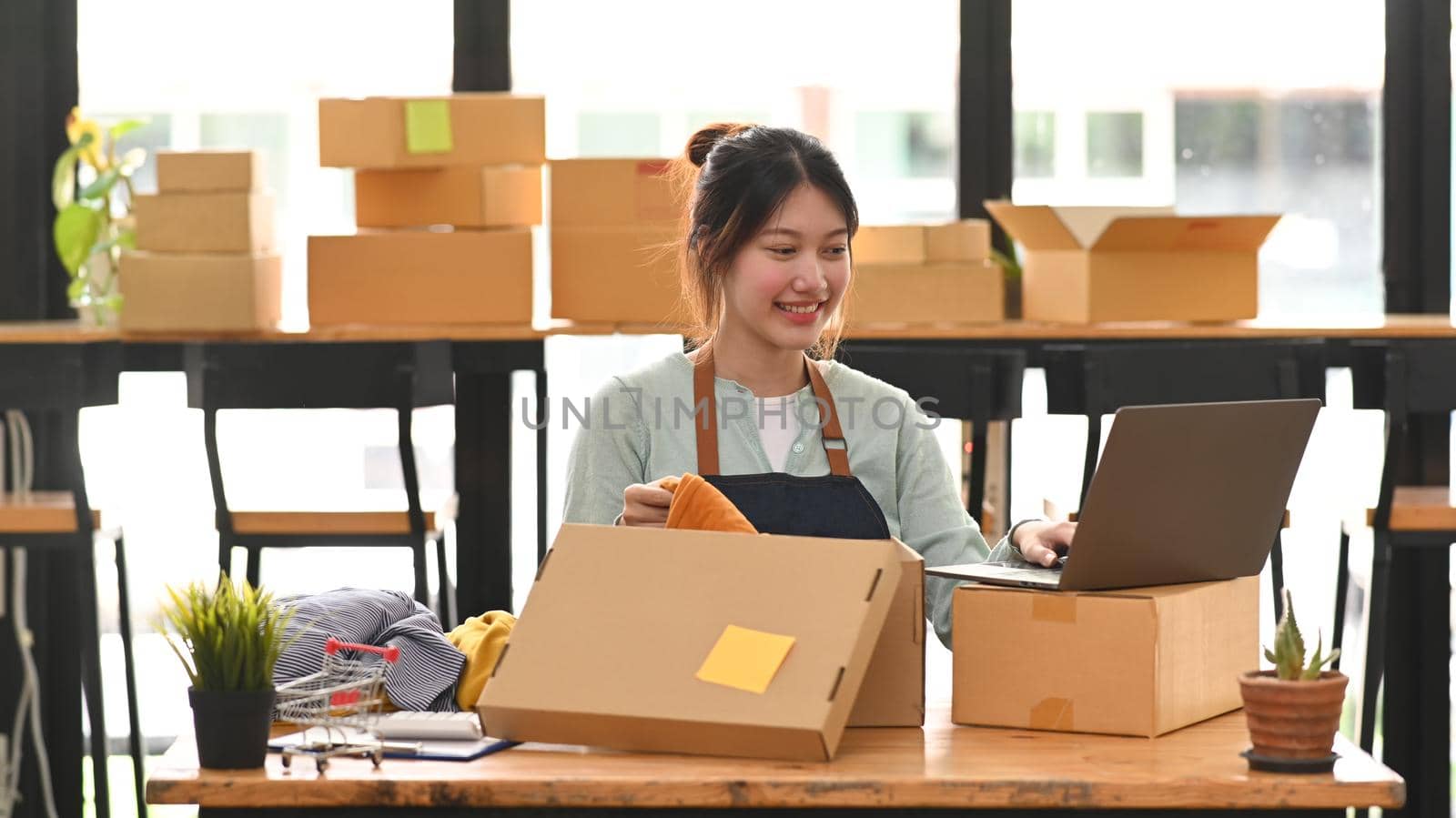 Smiling female small business owner using laptop during packing product in cardboard box for delivery to customers. Online selling, e-commerce.