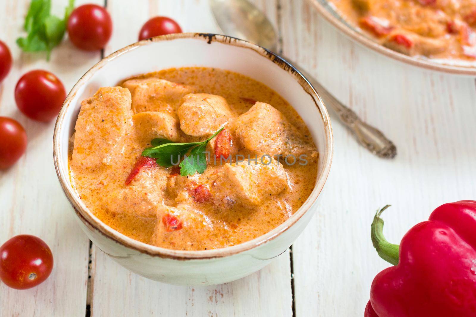 Delicious chicken stew with paprika in a bowl on a white wooden table. With fresh cherry tomatoes, red bell pepper and parsley. Traditional hungarian dish paprikash. Comfort food. Selective focus