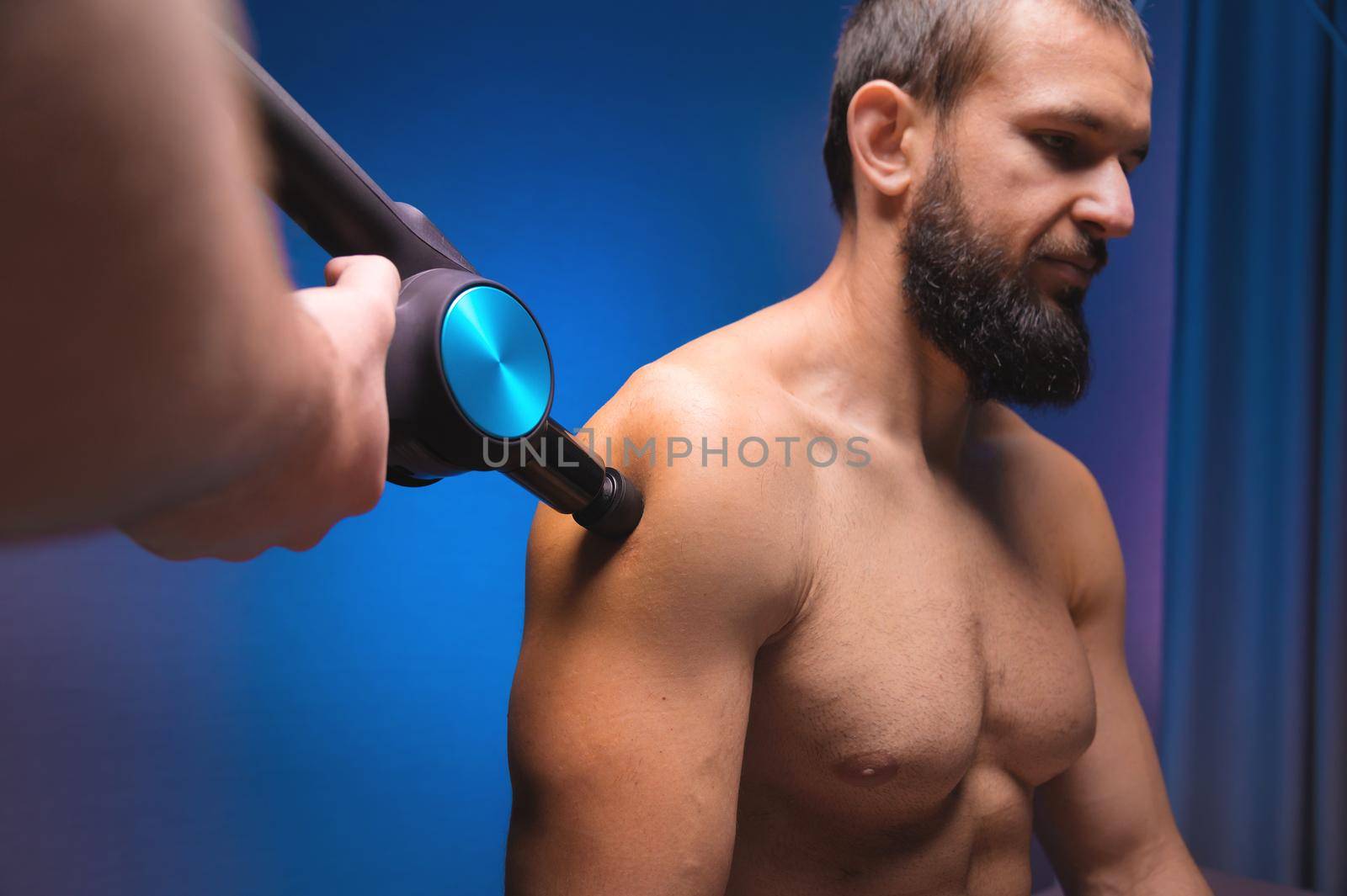 Medium shot of caucasian professional male massage therapist getting shoulder muscles with massage gun impact tool of muscular athlete, in spa treatments, sitting on massage table. by yanik88