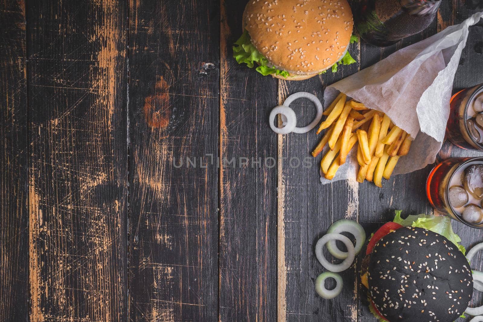 Set of black burgers with meat patty, cheese, tomatoes, mayonnaise, french fries and glass of cold cola soda with ice from above. Dark wooden rustic background. Space for text. Top view. Toned image