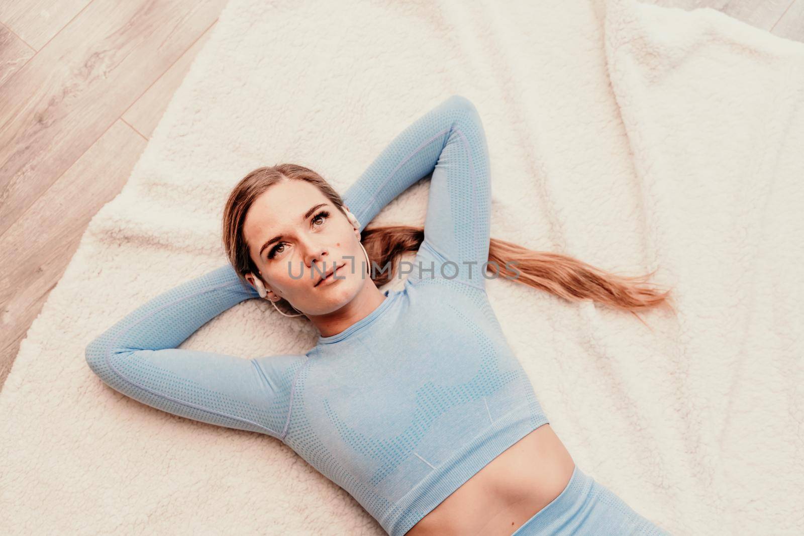 Top view portrait of relaxed woman listening to music with headphones lying on carpet at home. She is dressed in a blue tracksuit. by Matiunina