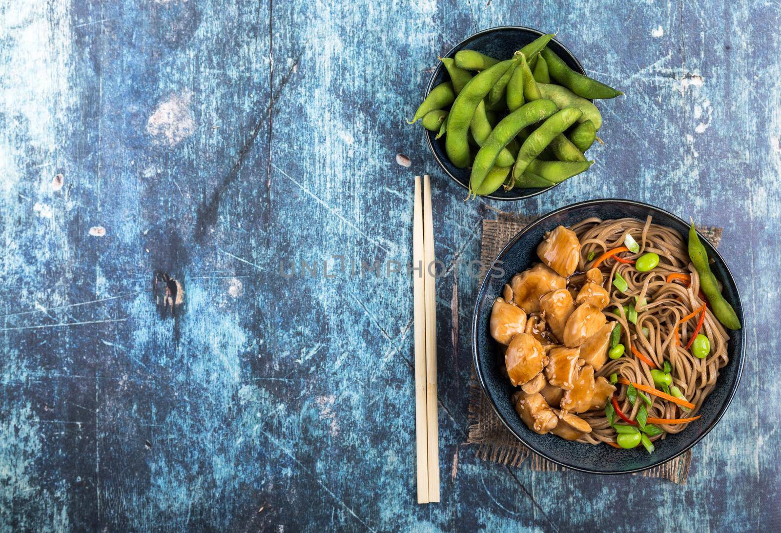 Asian noodles with chicken, vegetables, bowl, rustic wooden blue background. Space for text. Top view. Soba noodles, teriyaki chicken, edamame, chopsticks. Asian style dinner. Chinese/Japanese noodles