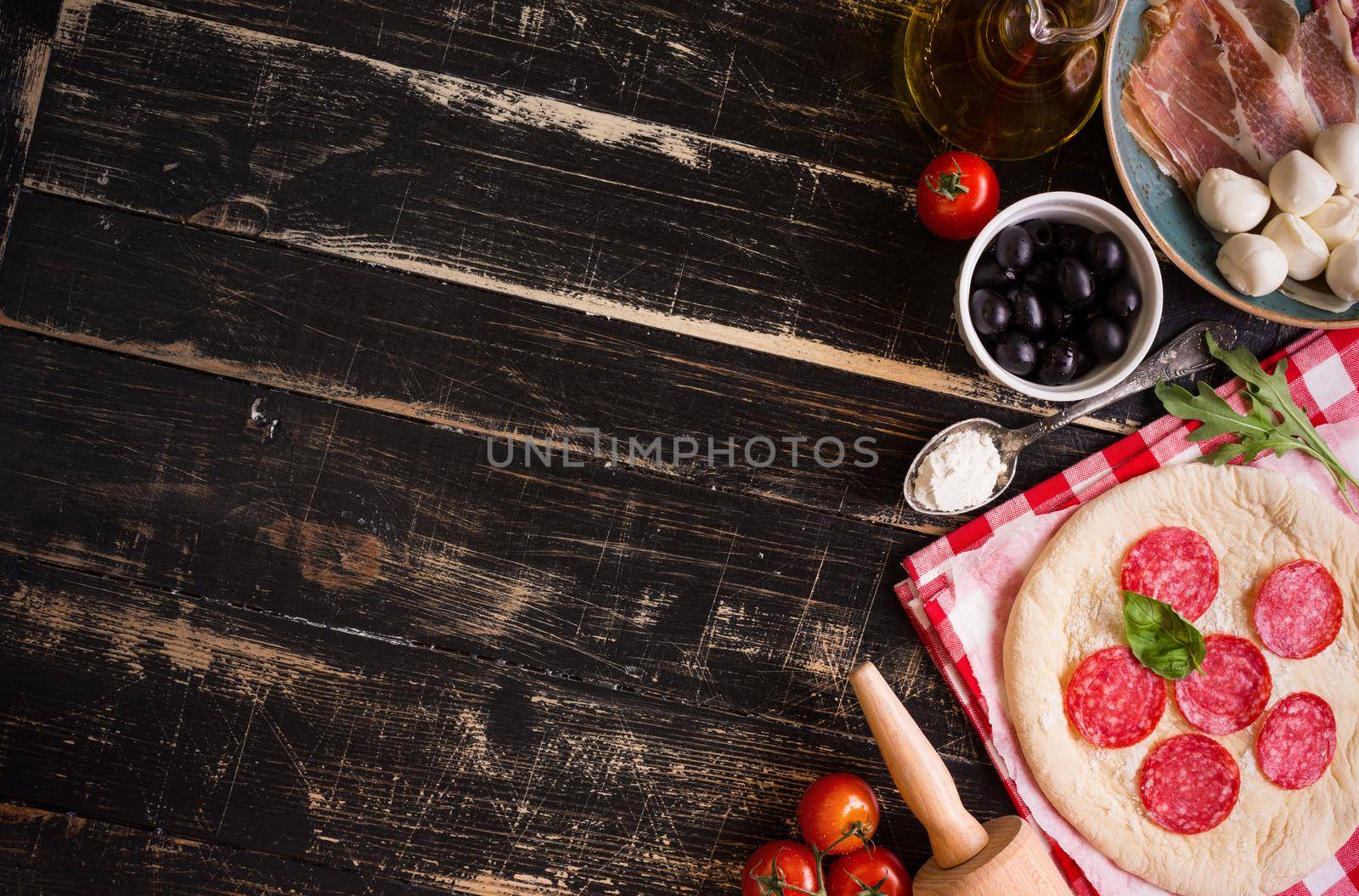 Pizza making background. Ingredients for making pizza. Space for text. Pizza dough, flour, cheese, mozzarella, tomatoes, basil, pepperoni, olives and rolling pin over black wooden background. Top view