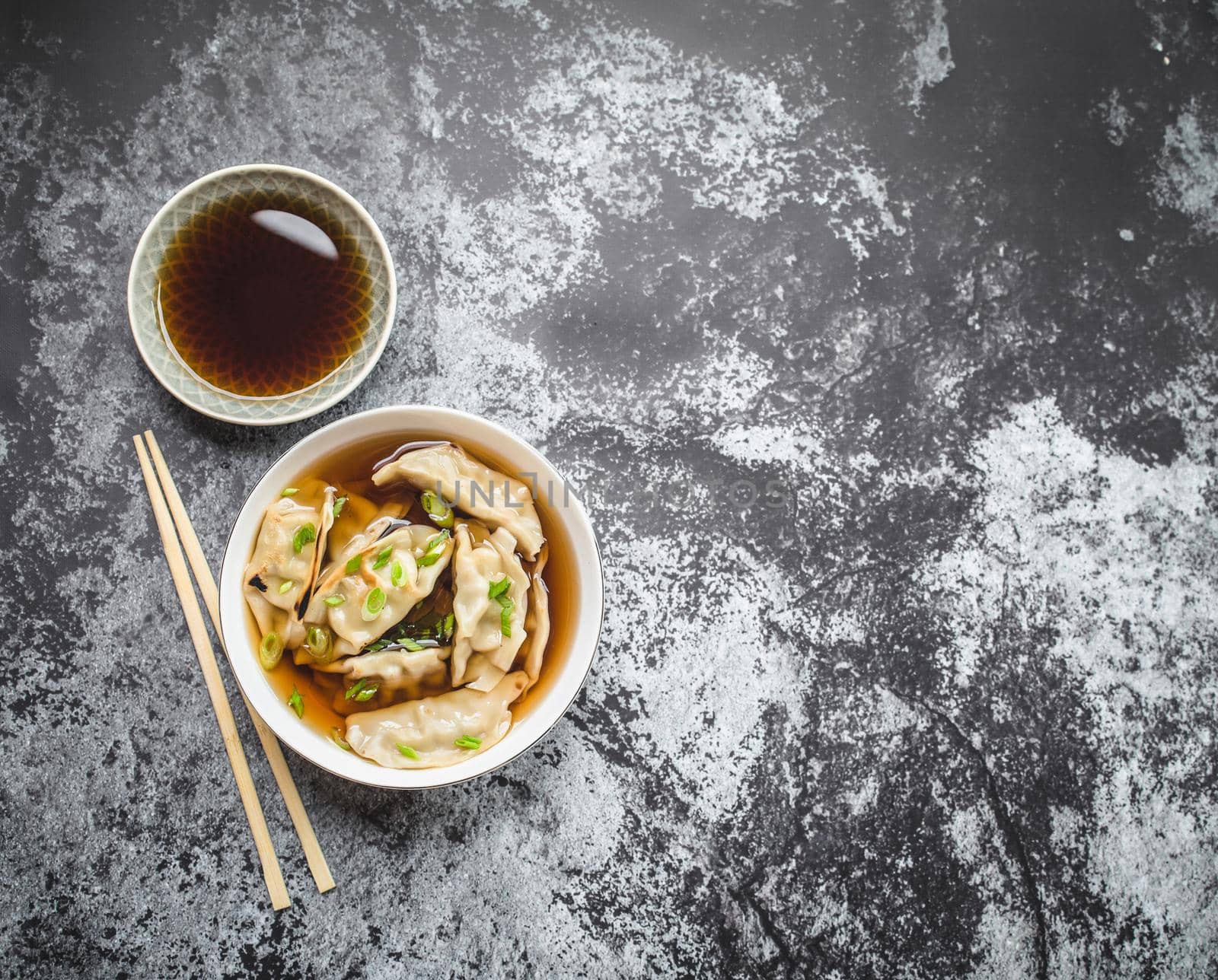 Asian dumplings in broth, bowl, chopsticks, soy sauce, rustic stone background. Top view. Space for text. Chinese dumplings for dinner. Closeup. Traditional Asian/Chinese cuisine. Overhead. Soup