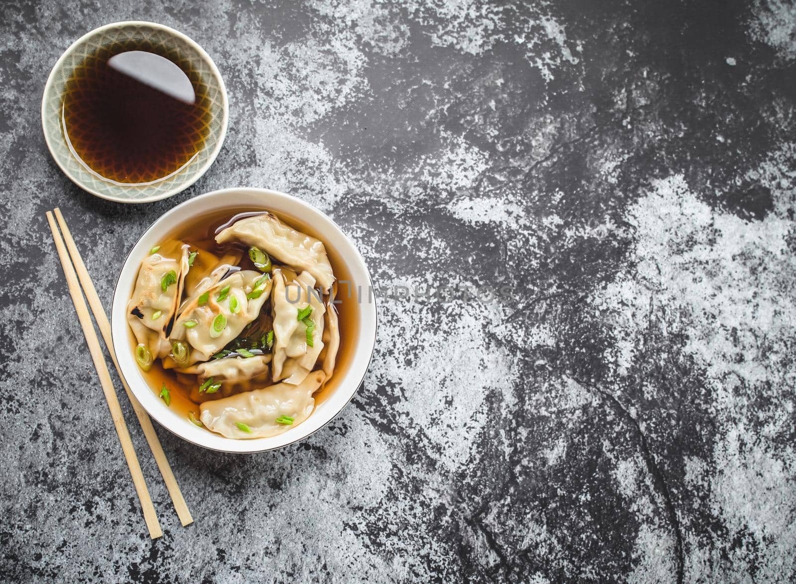 Asian dumplings in broth, bowl, chopsticks, soy sauce, rustic stone background. Top view. Space for text. Chinese dumplings for dinner. Closeup. Traditional Asian/Chinese cuisine. Overhead. Soup