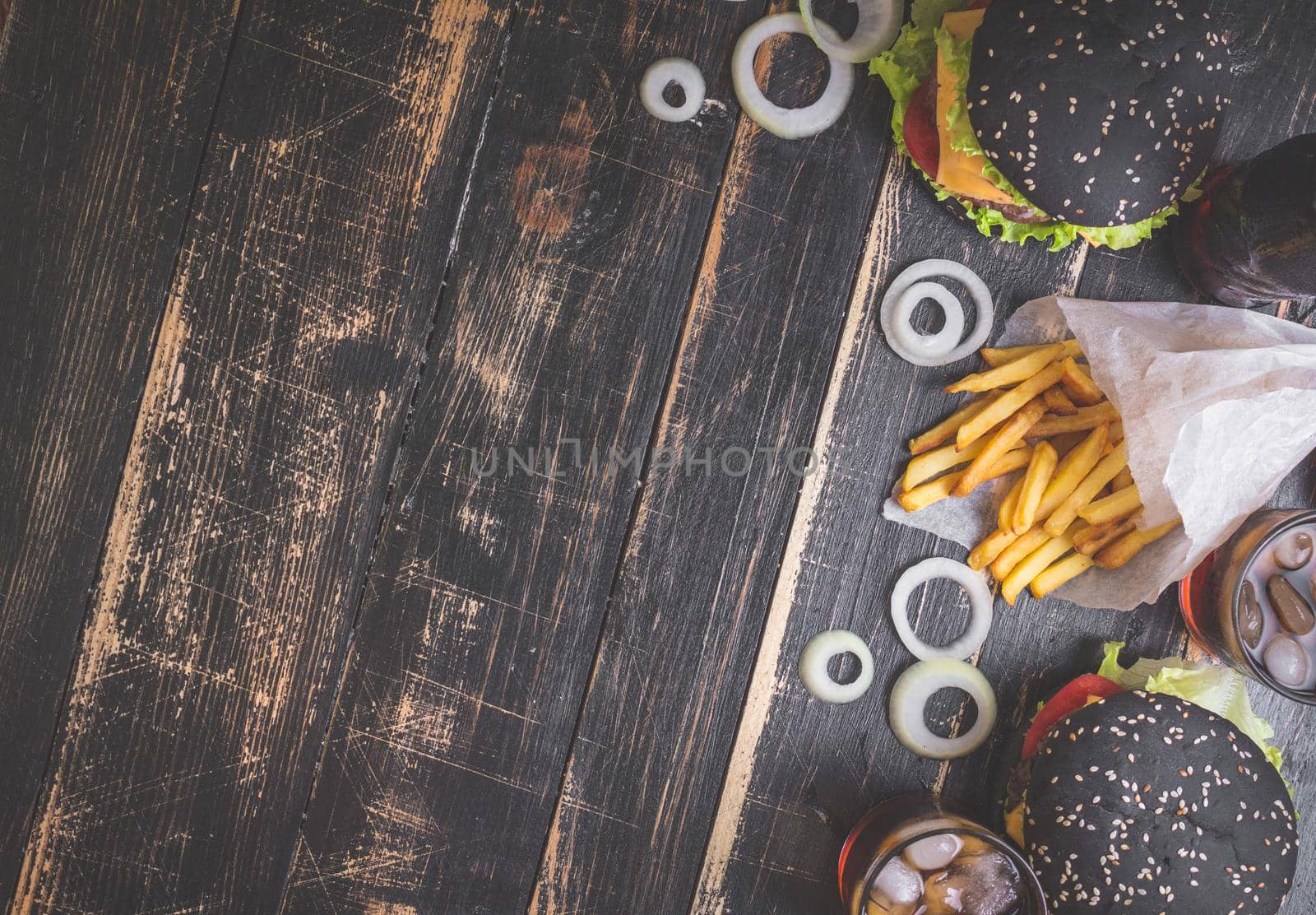 Set of black burgers with meat patty, cheese, tomatoes, mayonnaise, french fries and glass of cold cola soda with ice from above. Dark wooden rustic background. Space for text. Top view. Toned image