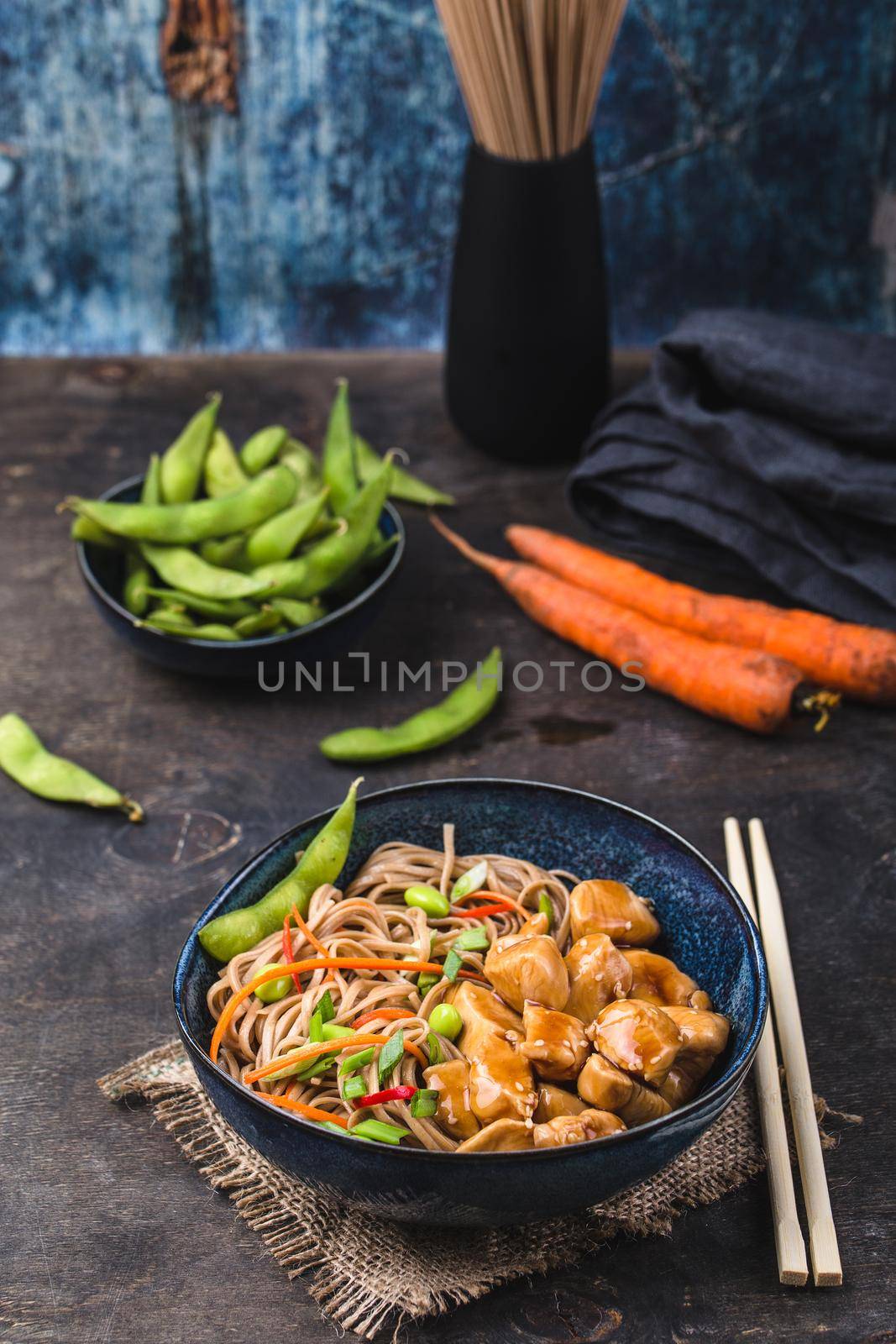 Asian noodles with chicken, vegetables in bowl, rustic wooden background. Soba noodles, teriyaki sauce chicken, edamame beans, sesame, chopsticks. Closeup. Asian style dinner. Chinese/Japanese noodles