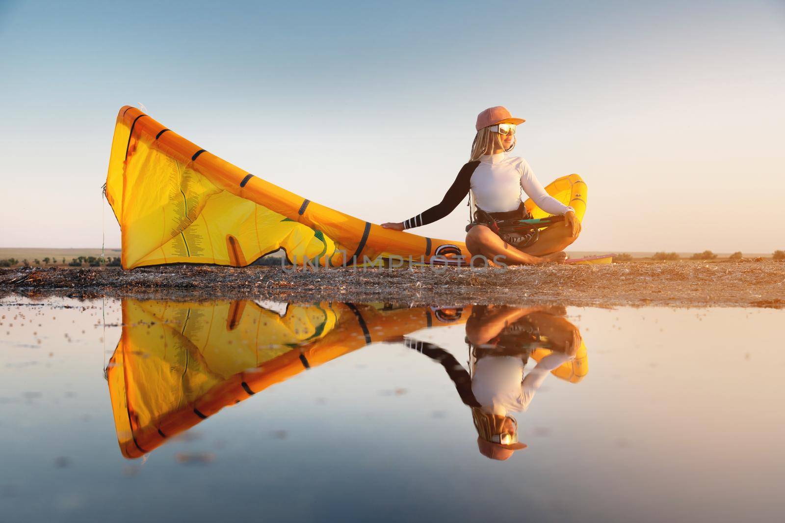 Attractive stylish young caucasian woman in cap sunglasses and kitesurfer outfit sits on the sandy shore next to her kite at sunset reflecting in the water.