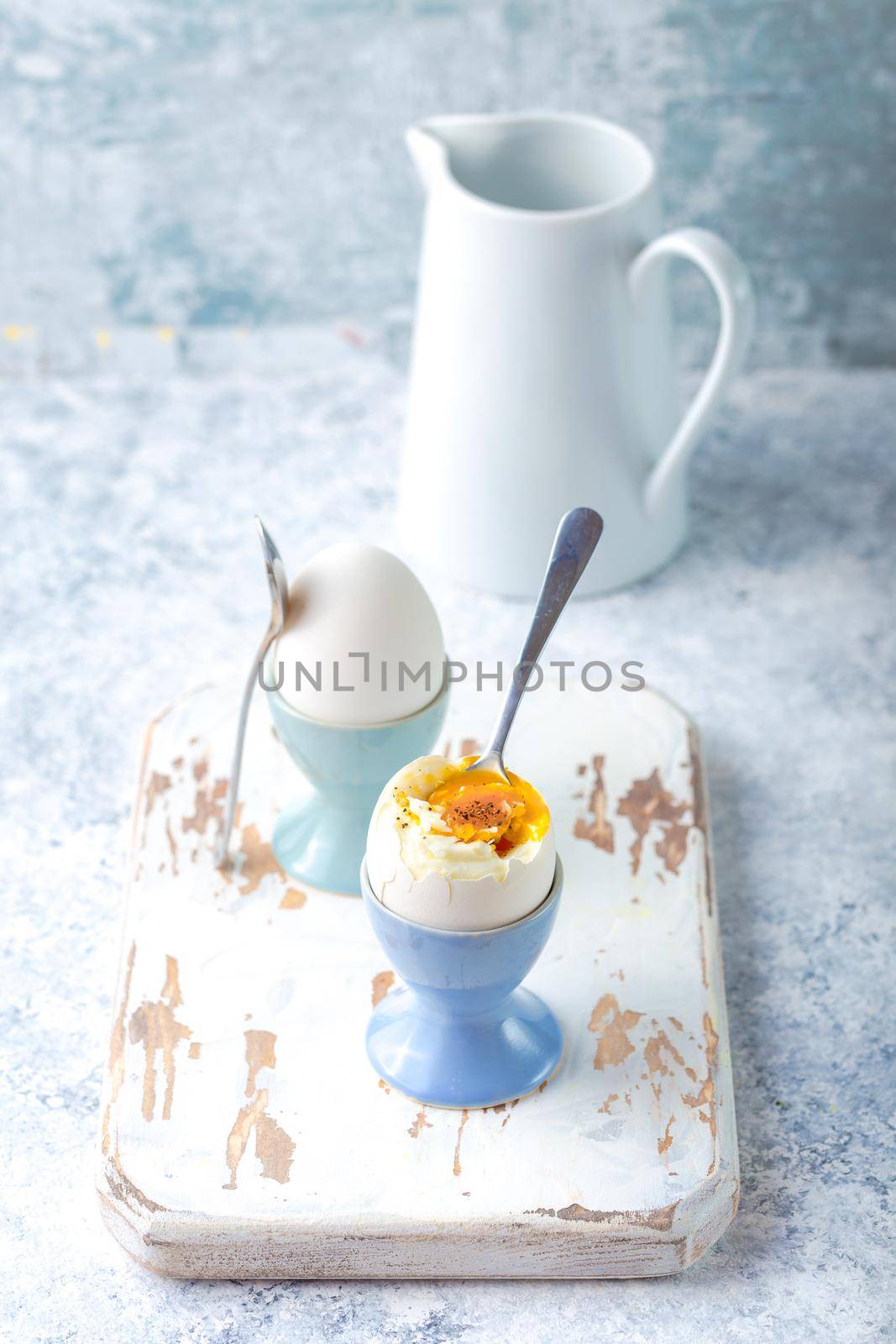 Fresh soft boiled eggs in stands, spoons, white wooden cooking board, white concrete rustic background. Soft eggs for healthy breakfast. Jar with milk. Selective focus. Egg protein fitness breakfast