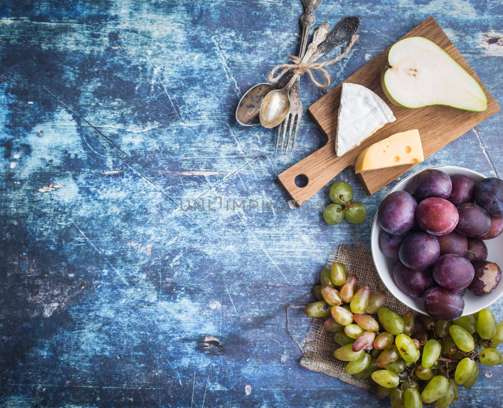 Fresh ripe pear, assorted cheeses platter, grapes, plums in bowl, blue wooden rustic background. Space for text. Top view. Fruits, Camembert, Emmental cheeses. Delicacy food. Snack/appetizer