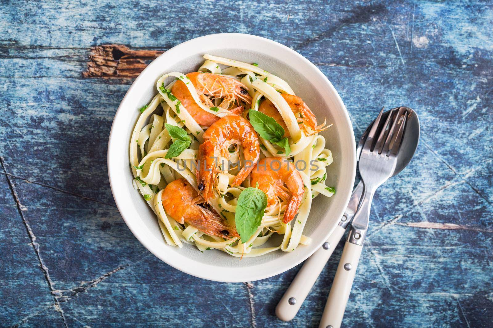 Homemade pasta with fried shrimps and basil on rustic wooden blue background. Pasta with seafood in plate. Top view. Close-up. Mediterranean cuisine. Pasta for dinner or lunch. Linguine with prawns