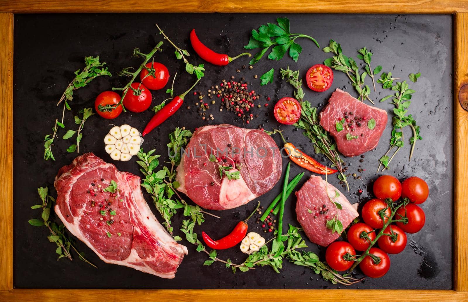 Different types of steaks set. Assorted raw meat on a black chalk board background with wood frame. Rib eye steak on the bone, veal shank (ossobuco), fillet with cherry tomatoes, hot pepper and herbs.