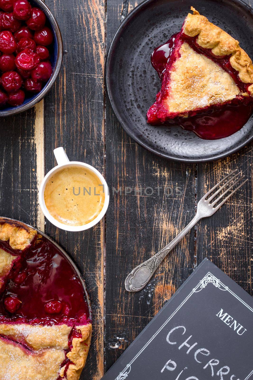 Homemade sliced cherry pie with flaky crust, cup of coffee, bowl with cherries and menu chalkboard on the black wooden table. Top view