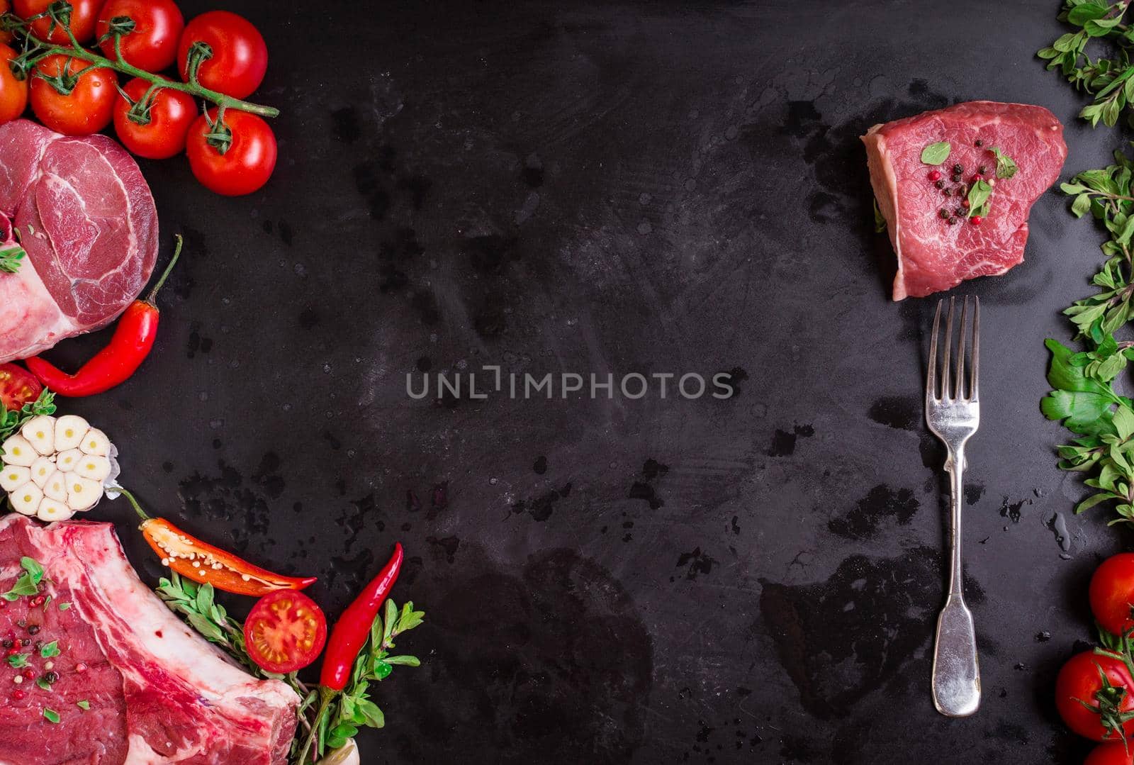 Raw juicy meat steaks ready for roasting on a black chalk board background. Meat on a fork. Veal shank (ossobuco), rib eye steak on the bone, fillet with cherry tomatoes, hot pepper and herbs.