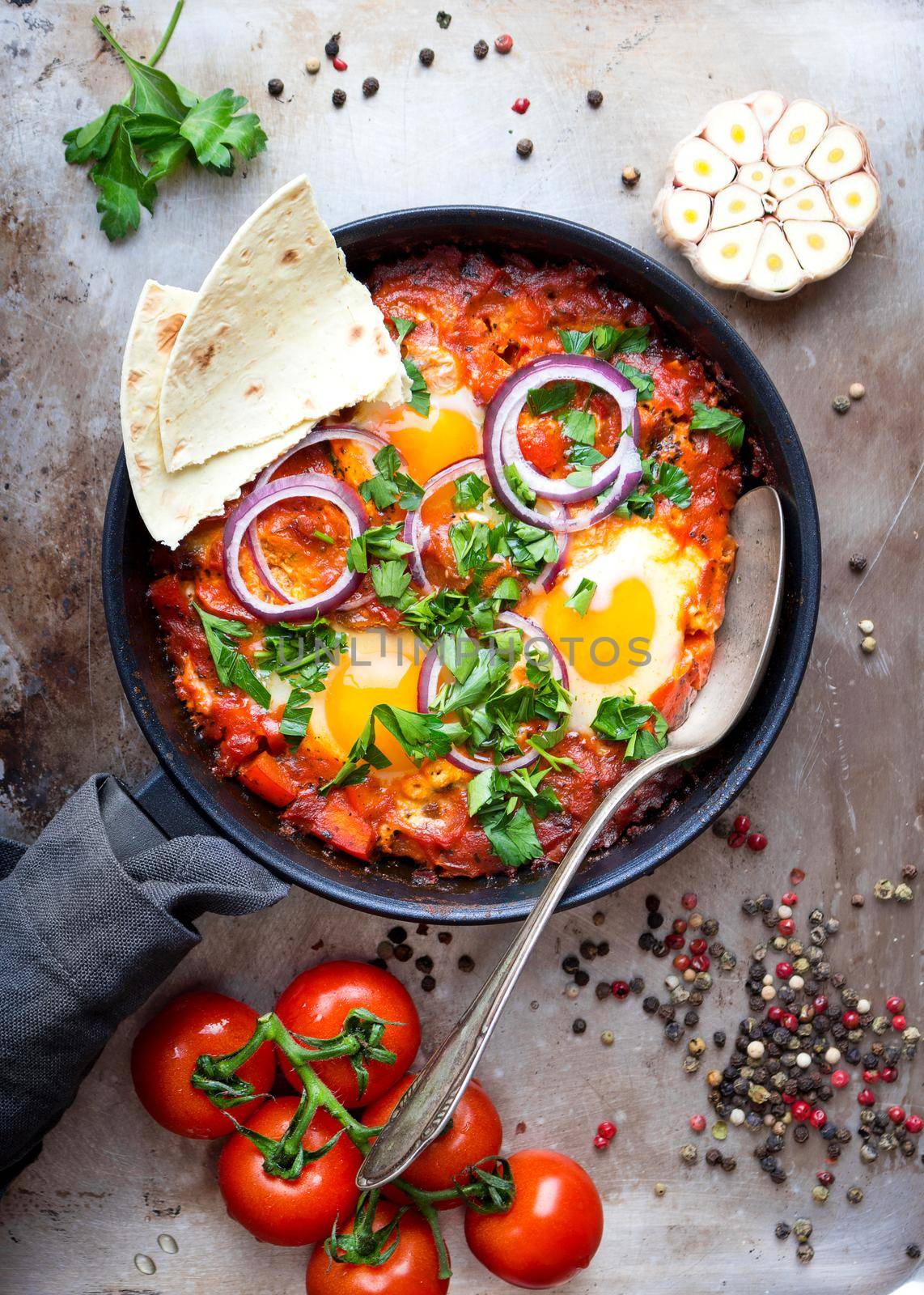 Shakshuka with pita bread in a pan. Middle eastern traditional dish. Fried eggs with tomatoes, bell pepper, vegetables and herbs. Shakshouka on a table. Top view. Sunny side up eggs. From above