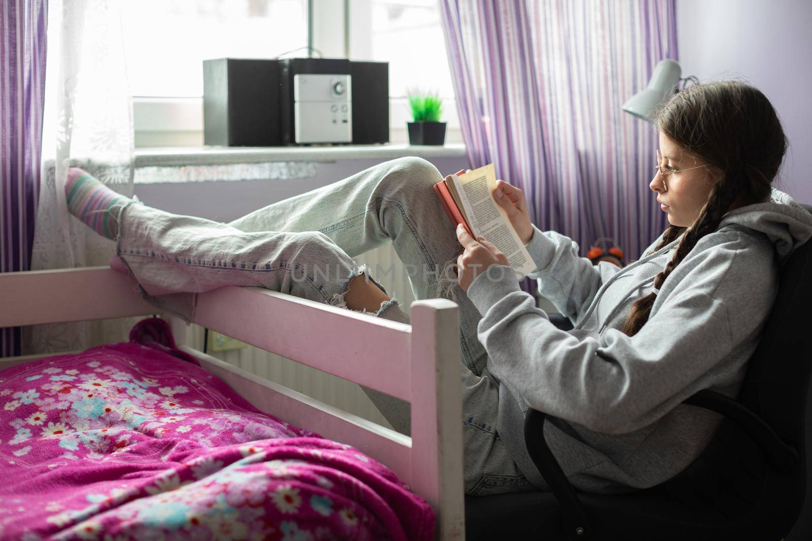 A girl is studying from a school book while lying in a strange position in her room. A young girl wearing glasses with braids. A lover of books.